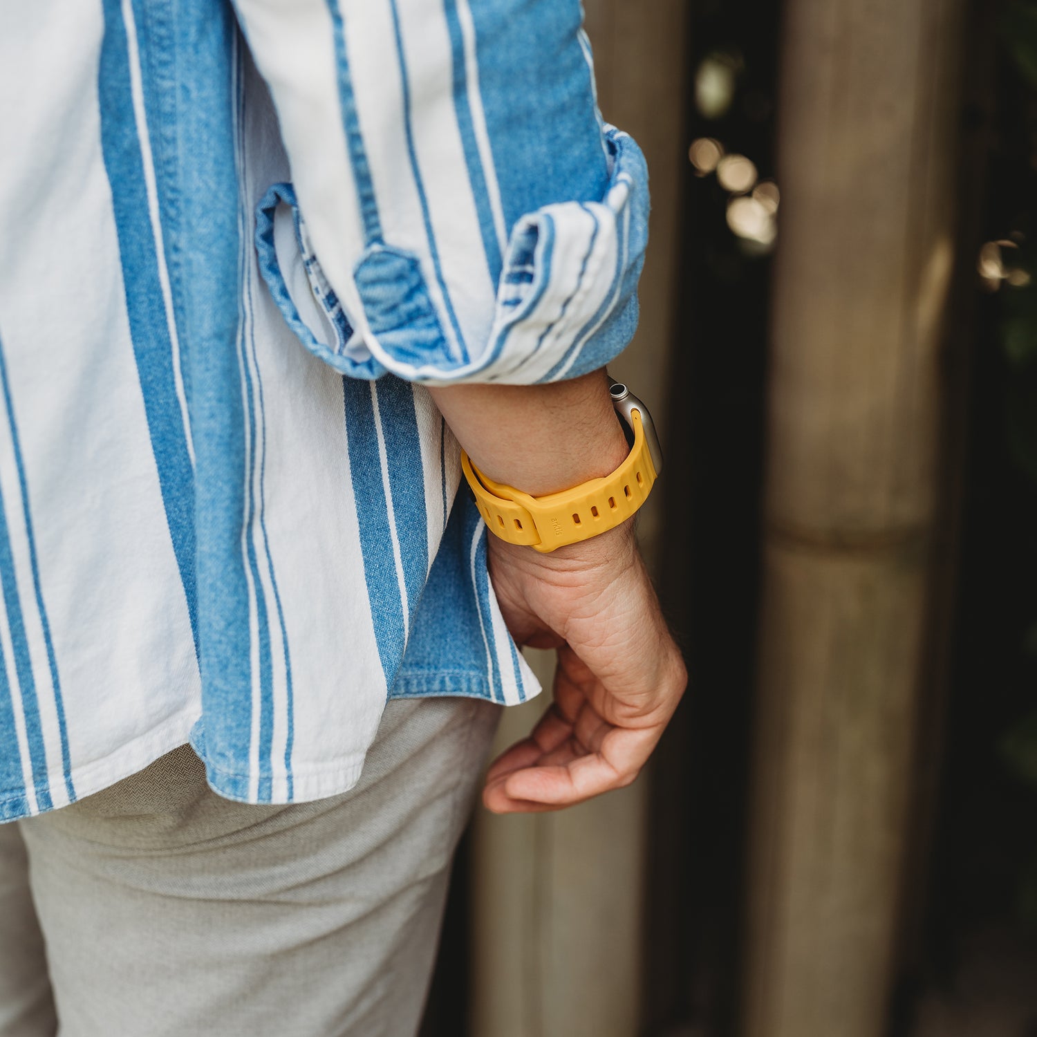 Eine Person in einem blau-weiß gestreiften Hemd und einer hellen Hose steht mit dem Rücken zur Kamera und trägt ein arktisband Apple Watch Silikonarmband „Active“ von arktisband am linken Handgelenk, die Hand hängt entspannt an der Seite.