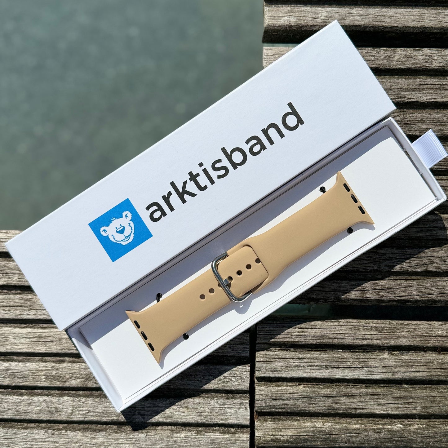 Das arktisband Apple Watch Silikonarmband „Aqua“ in Beige mit silberner Schnalle wird in einer weißen Box mit blauem Eisbär-Logo auf Holz präsentiert – passend für aktive Tage am Wasser.