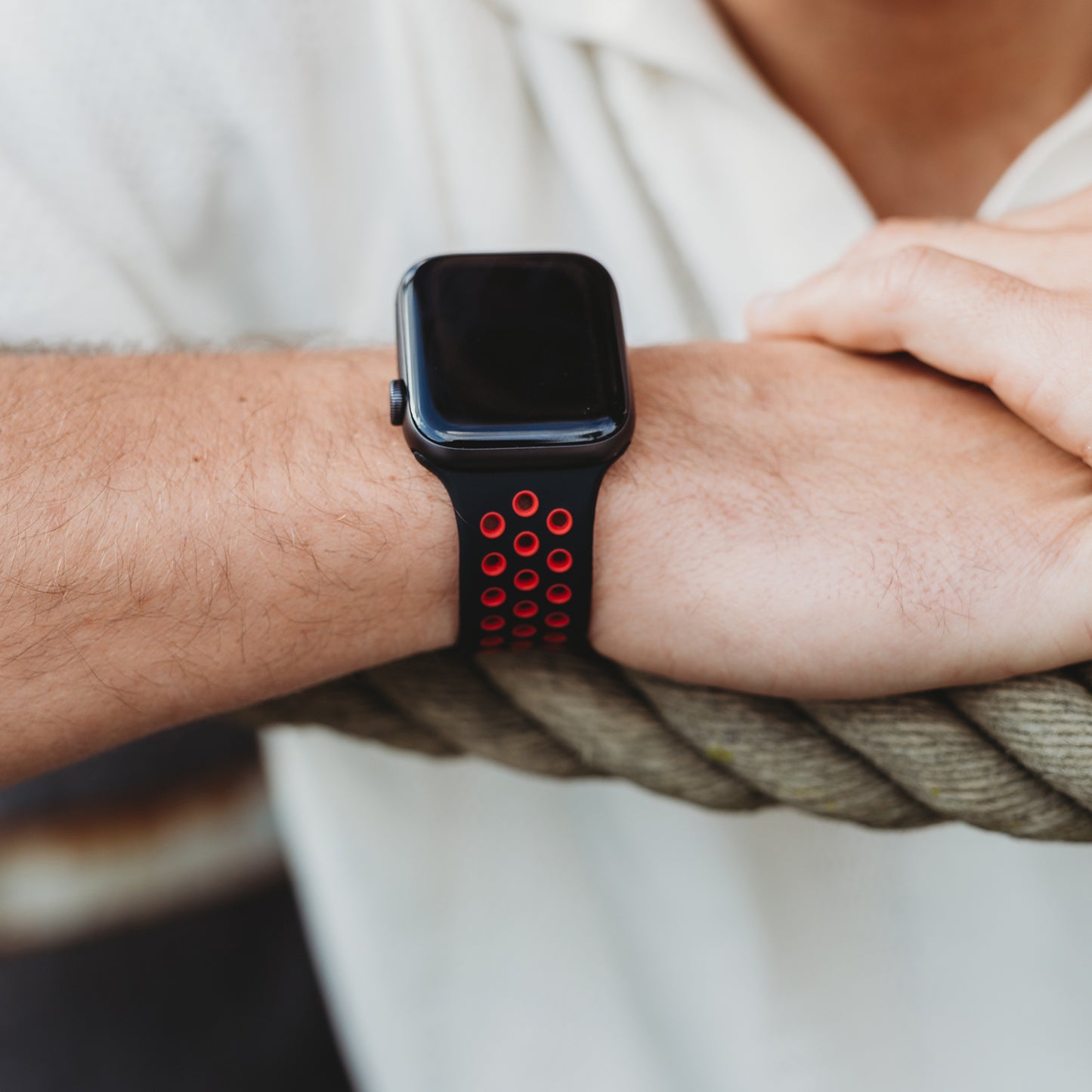 Der Unterarm einer Person ruht auf einem dicken Seil. Sie trägt das arktisband Apple Watch Silikonarmband "Energy" in Schwarz und Rot. Das Smartwatch-Armband von arktisband ist mit der Apple Watch kompatibel. Die Person trägt ein weißes Kurzarmhemd.