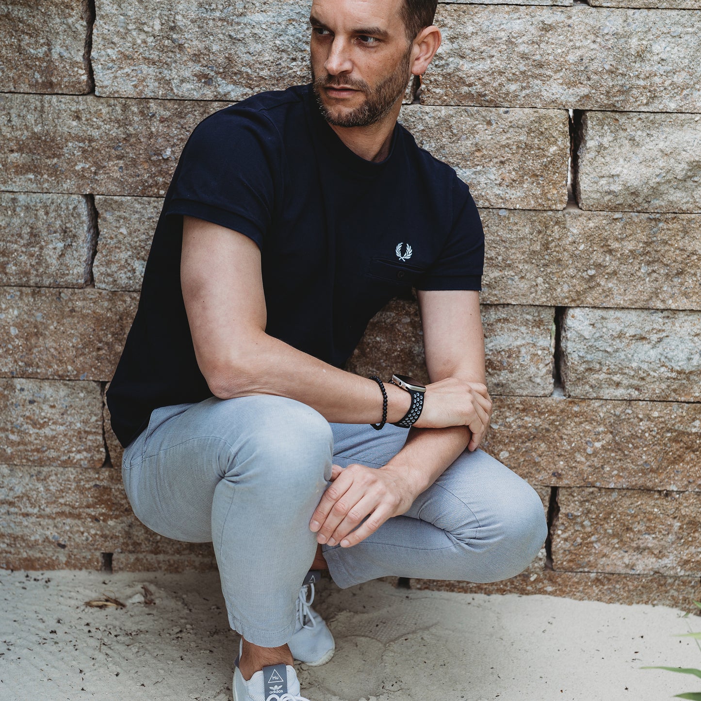Ein Mann mit kurzen Haaren und Bart kauert an einer Steinmauer. Er trägt ein schwarzes T-Shirt, eine hellgraue Hose, Turnschuhe und ein Arktisband Apple Watch Silikonarmband „Energy“ am linken Handgelenk und blickt nach links.