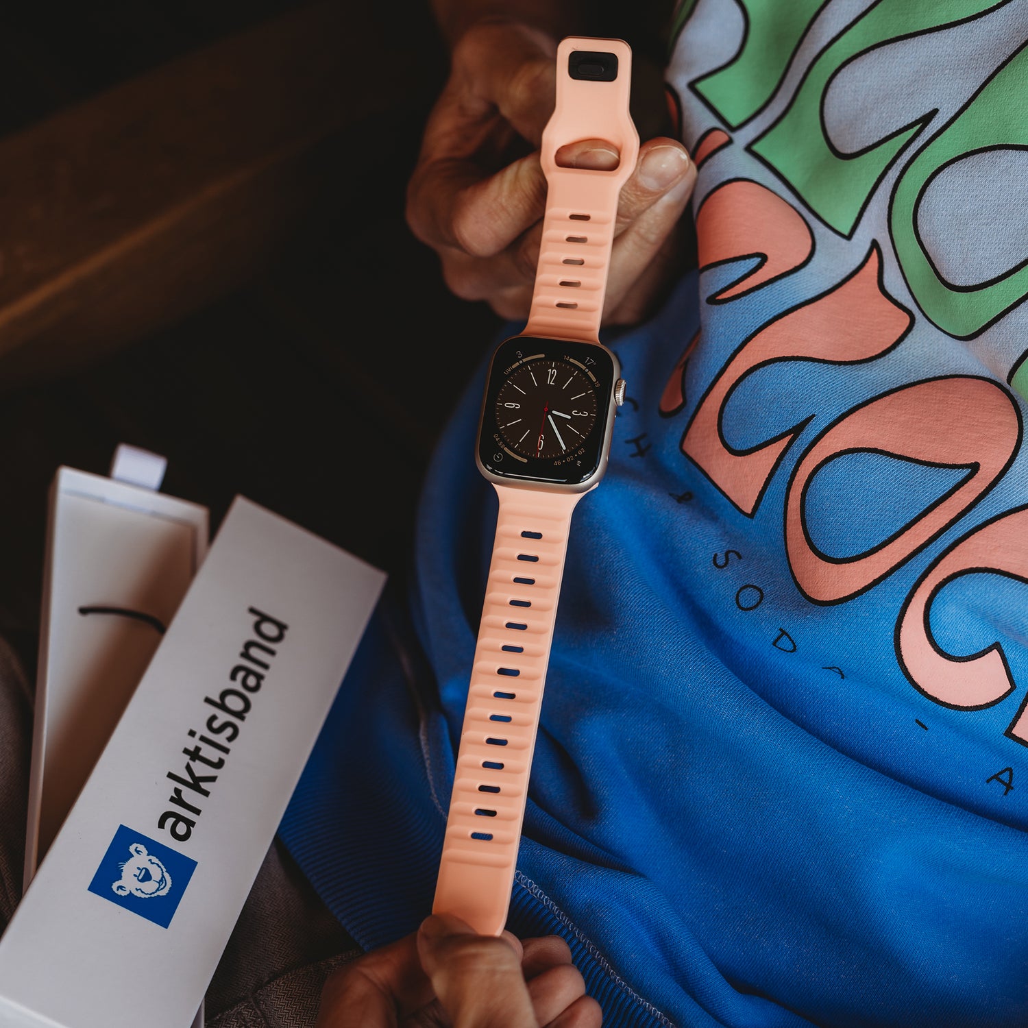 Eine Person hält eine Apple Watch mit dem Arktisband Apple Watch Silikonarmband „Active“ in Pfirsichfarbe über ihrem Schoß und trägt ein blaugrünes T-Shirt mit Grafik. Daneben steht eine offene Arktisband-Box für einen sportlichen Look.