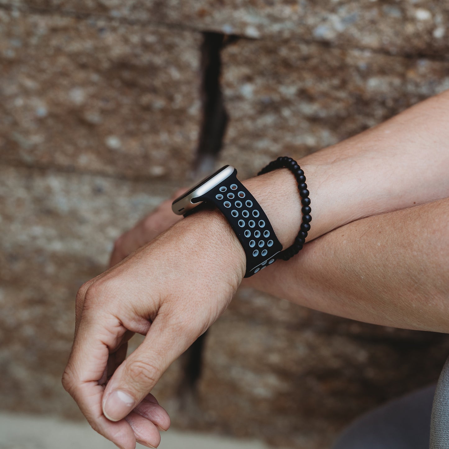 An einem linken Handgelenk trägt man das arktisband Apple Watch Silikonarmband „Energy“ von arktisband, ein schwarzes Band mit mehreren runden Löchern, das neben einem schwarzen Perlenarmband vor einem rauen Steinhintergrund abgebildet ist.