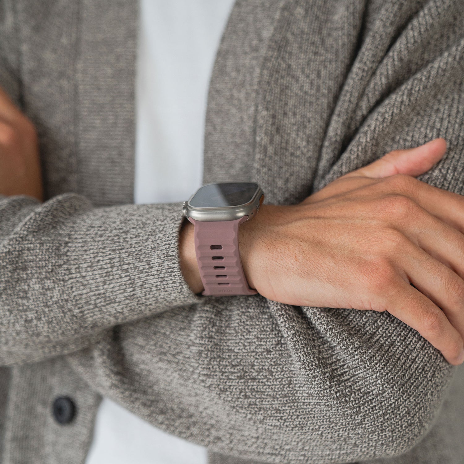 Eine Person in einer grauen Strickjacke und einem weißen Hemd trägt mit verschränkten Armen ein arktisband Apple Watch Silikonarmband „Active“ mit atmungsaktivem Design und rosa Finish von arktisband für ganztägigen Tragekomfort.
