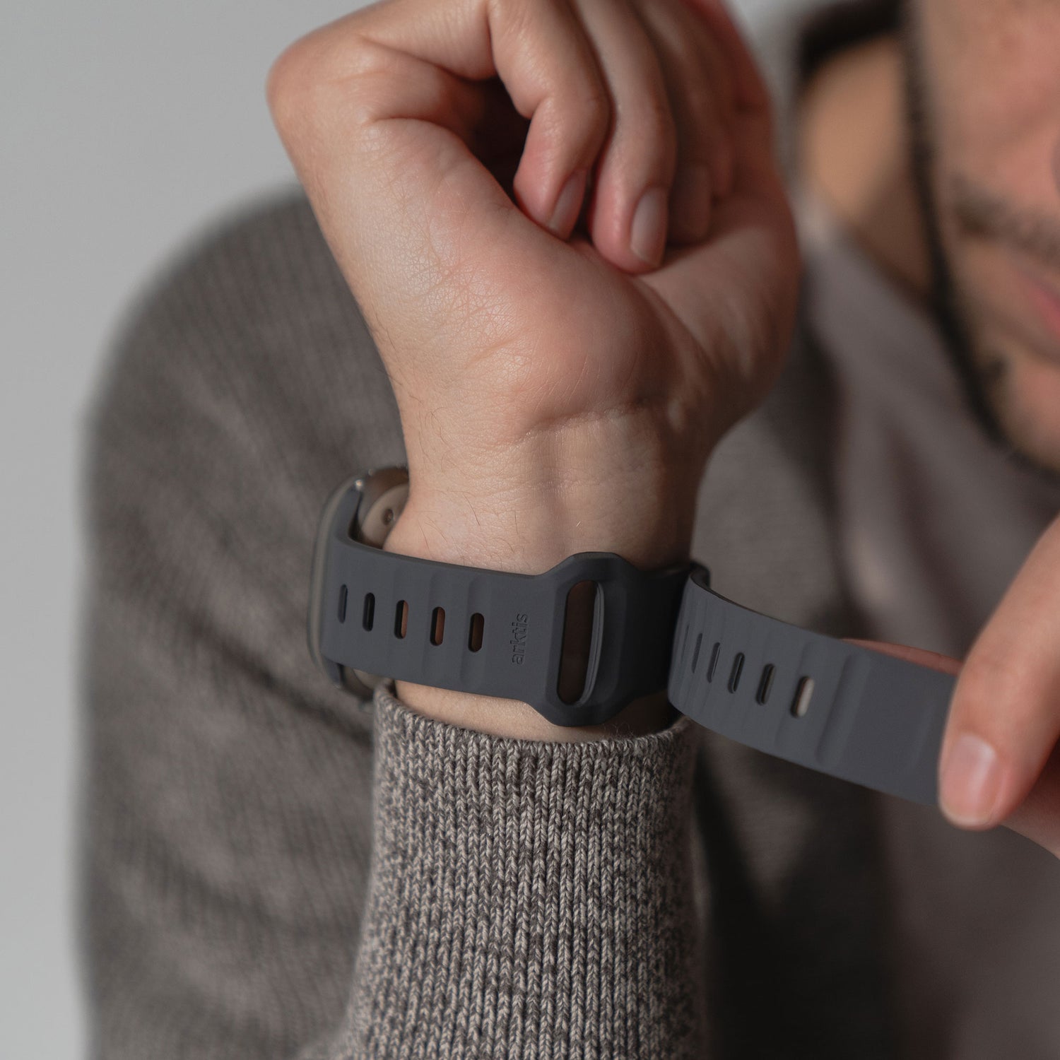 Eine Person befestigt das schwarze arktisband Apple Watch Silikonarmband „Active“ von arktisband am Handgelenk. Mit einem grauen Strickpullover trägt sie einen sportlichen Look, wobei ihr Gesicht teilweise aus dem Bild verschwindet.