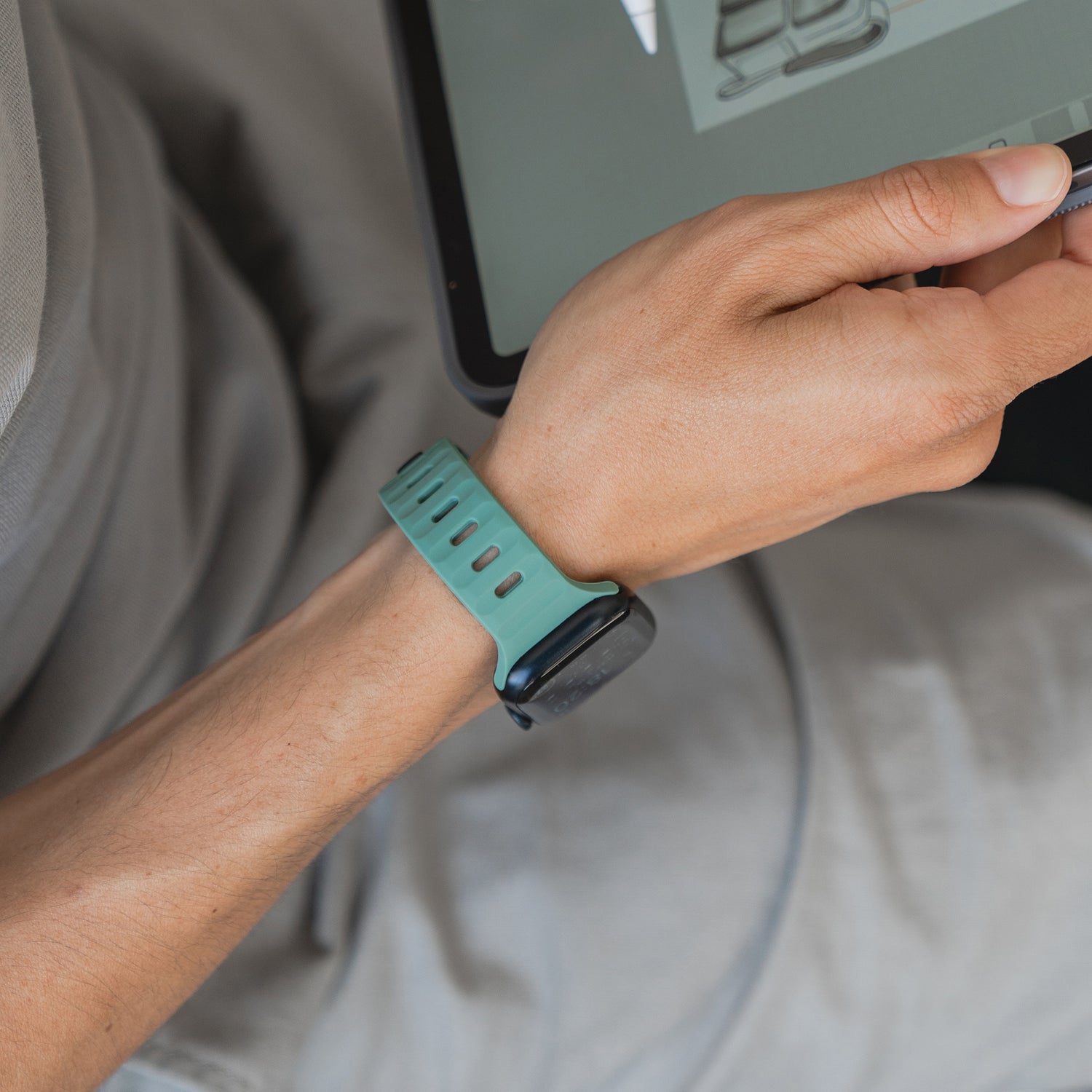 Eine Person trägt ein grünes Arktisband Apple Watch Silikonarmband „Active“ und hält einen Stift, während sie ein Tablet benutzt. Die Person ist hell gekleidet und sitzt bequem.