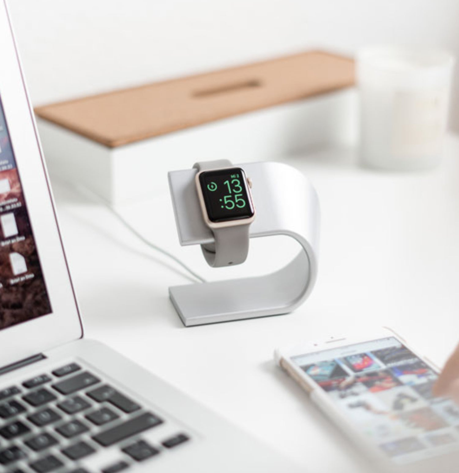 Eine Smartwatch auf einem silbernen Ständer neben einem Laptop und einer Person, die ein Smartphone auf einem weißen Schreibtisch benutzt.