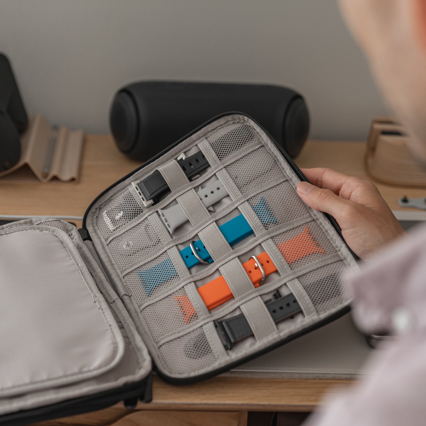 Eine Person hält eine offene Künstlerband Kleine Reisetasche für Apple Watch Armbänder & Zubehör mit Netzfächern, die mehrere Armbänder in Blau, Orange und Schwarz enthält, über einem Schreibtisch.