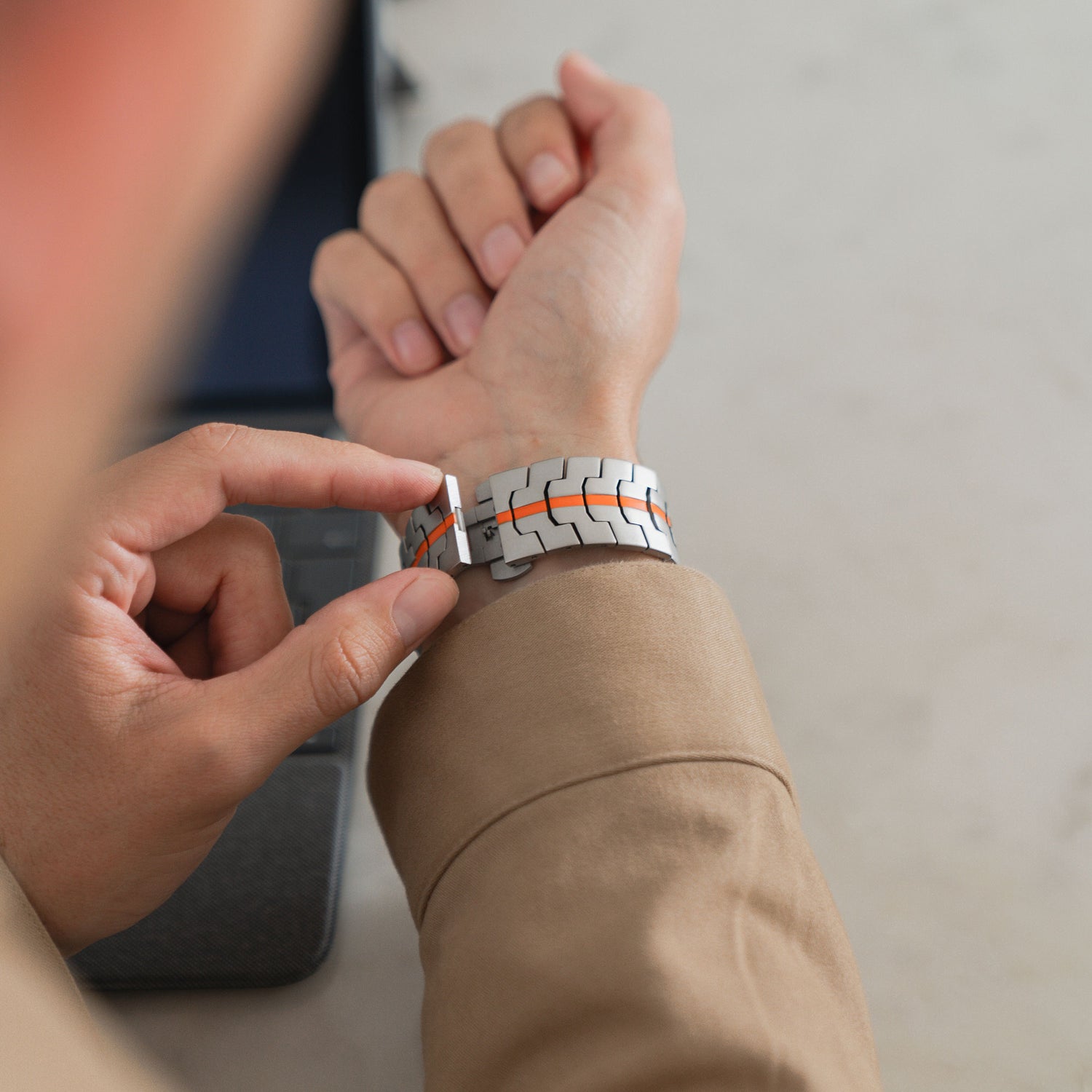 Eine Person in einer beigen Jacke befestigt ein arktisband Apple Watch Ultra Armband TITANIUM F-15 von arktisband mit orangefarbenen Akzenten an ihrem linken Handgelenk in der Nähe eines Laptops auf einer hellen Oberfläche.