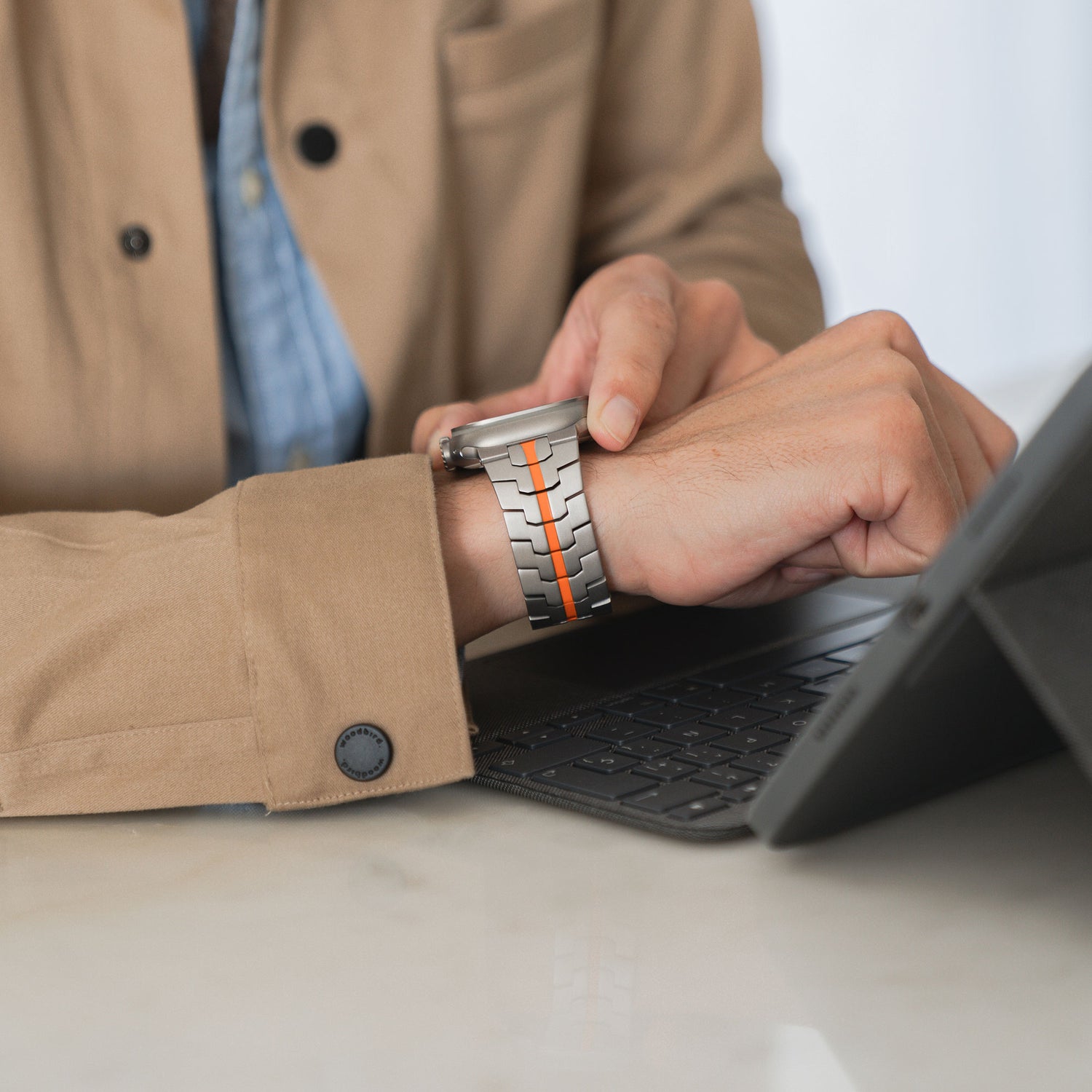 Eine Person, die eine beige Jacke und ein Arktisband Apple Watch Ultra Armband TITANIUM F-15 trägt, verwendet ein Tablet mit einer Tastaturhülle auf einer weißen Oberfläche und stellt die Uhr an ihrem Handgelenk ein.