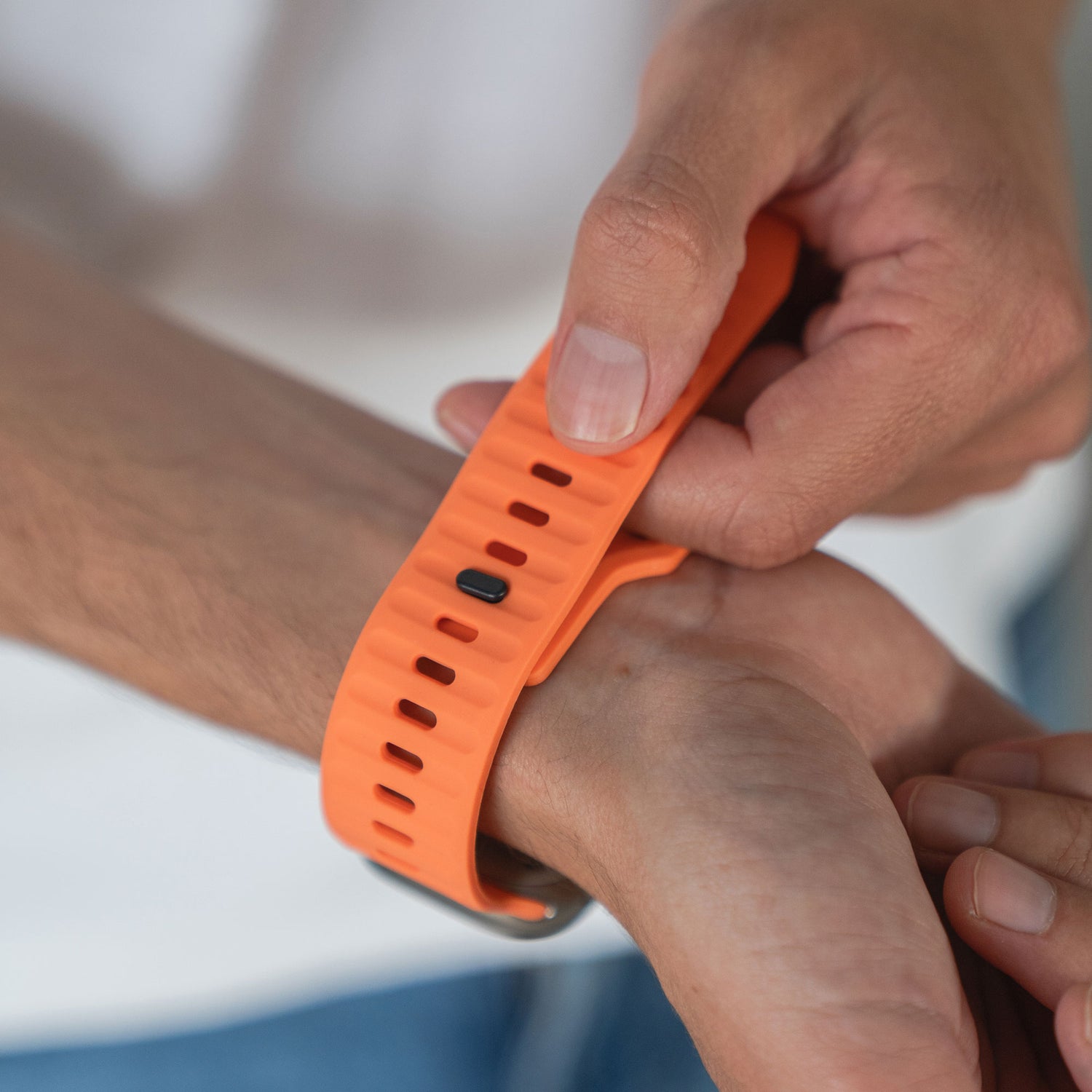 Jemand befestigt ein arktisband Apple Watch Silikonarmband „Active“ mit mehreren Löchern und einer kleinen schwarzen Schließe um sein Handgelenk und passt das orangefarbene Band von arktisband für optimale Atmungsaktivität an. Der Hintergrund ist unscharf.