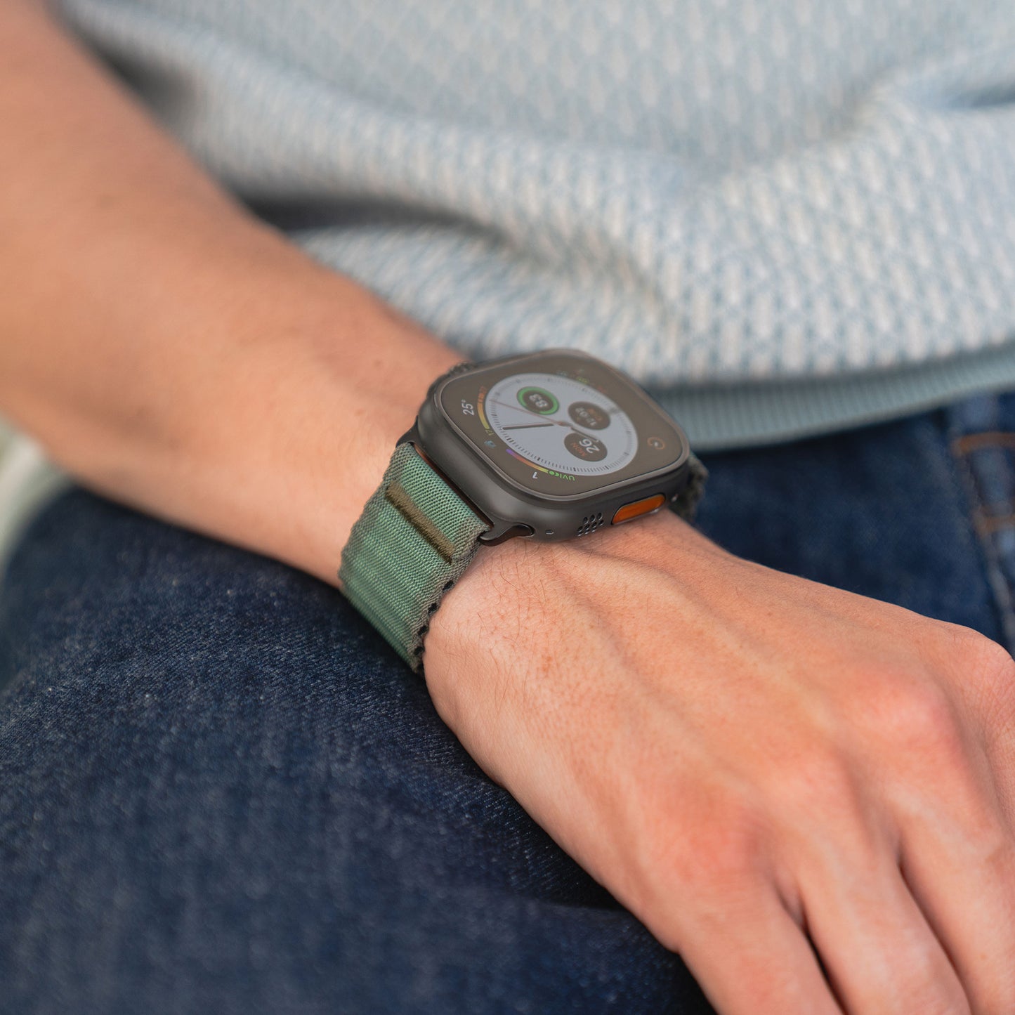 Eine Person trägt das grüne Arktisband Apple Watch Nylonarmband „Venture“, kombiniert mit Blue Jeans und einem hellblauen gemusterten Oberteil. Die Apple Watch zeigt App-Symbole und Fitnessdaten an – perfekt für Outdoor-Fans.
