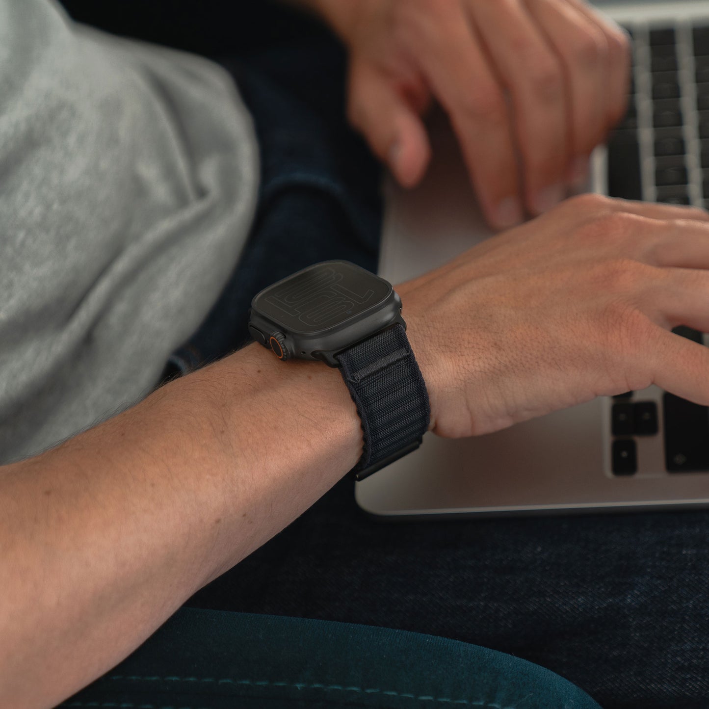 Jemand trägt ein Arktisband Apple Watch Nylonarmband „Venture“ von Arktisband und tippt auf einem Laptop. Nur seine Hände, ein Teil seines Arms und der Laptop sind sichtbar. Er trägt ein hellgraues Hemd und dunkle Jeans.