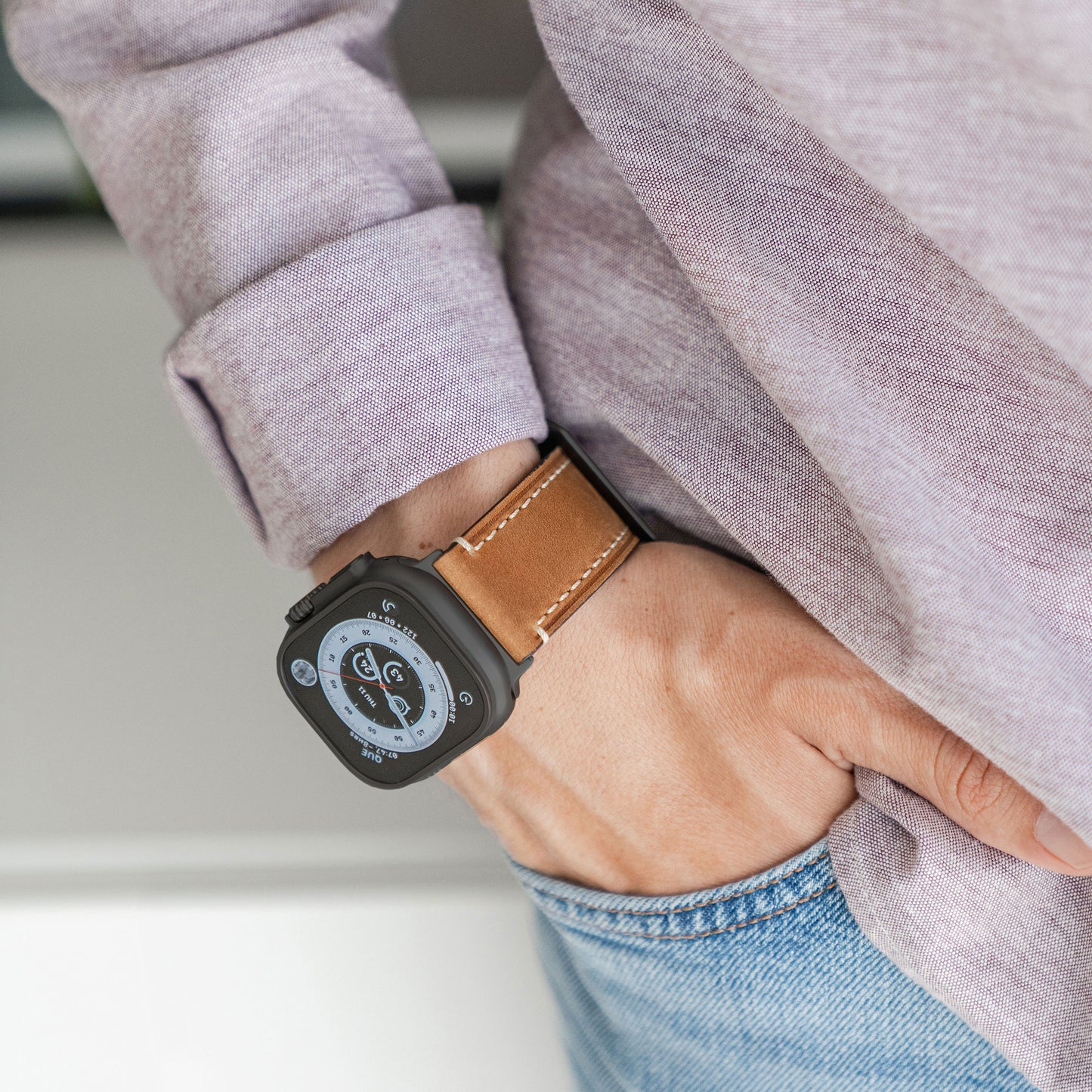Eine Person in einem beigen Langarmhemd und Blue Jeans hat ihre linke Hand in der Tasche und zeigt eine Apple Watch Ultra mit einem runden Zifferblatt im Analogstil und einem Arktisband PALERMO GRANDE Lederarmband.