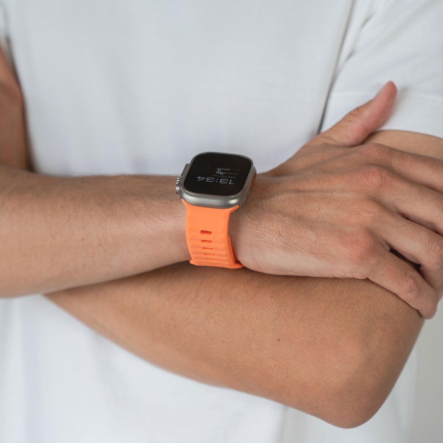 Eine Person im weißen T-Shirt verschränkt die Arme und präsentiert ein arktisband Apple Watch Silikonarmband „Active“ in leuchtendem Orange – ein sportlicher Look. Die Uhr zeigt die Uhrzeit 13:34 Uhr an.