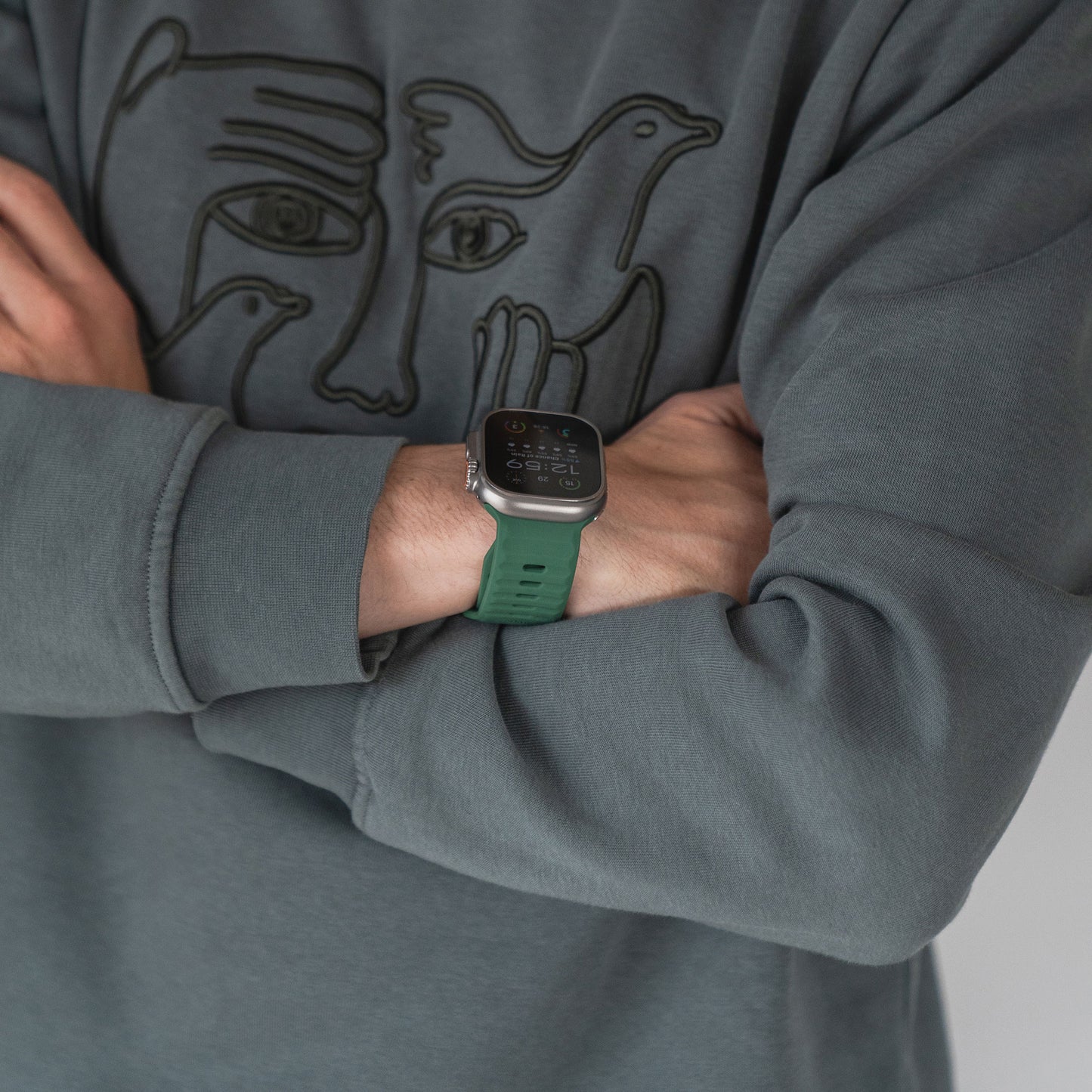 Eine Person in einem grauen Sweatshirt mit abstraktem Gesichts- und Vogeldesign steht mit verschränkten Armen da und zeigt eine Smartwatch mit dem Arktisband Apple Watch Silikonarmband „Active“ mit atmungsaktivem Design am Handgelenk.