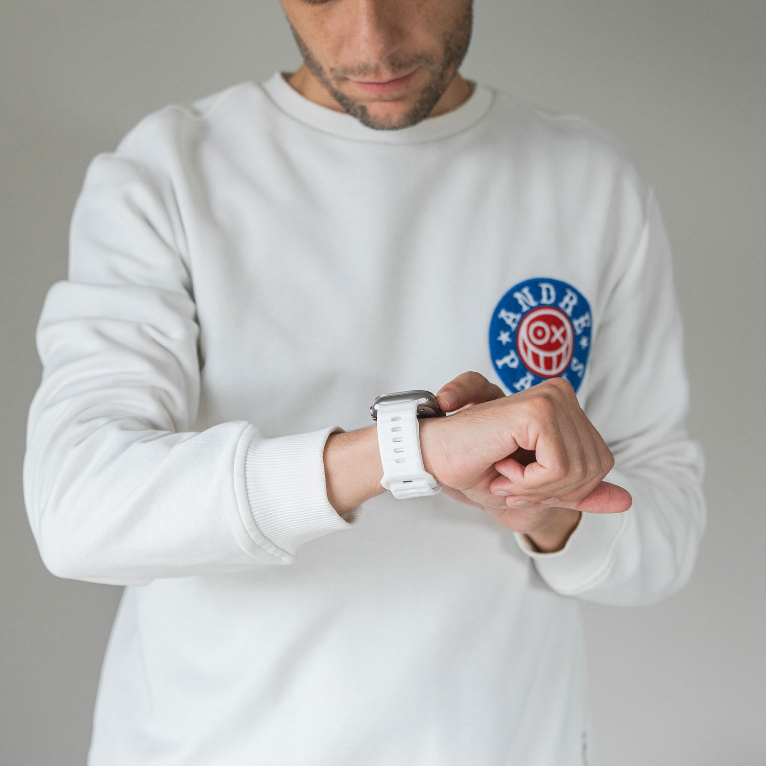 Eine Person in einem weißen Sweatshirt mit blau-rotem Patch blickt auf ihre Apple Watch, die mit einem arktisband Apple Watch Silikonarmband „Active“ von arktisband ausgestattet ist, und berührt die Uhr mit der rechten Hand. Der Hintergrund ist schlicht und hell.