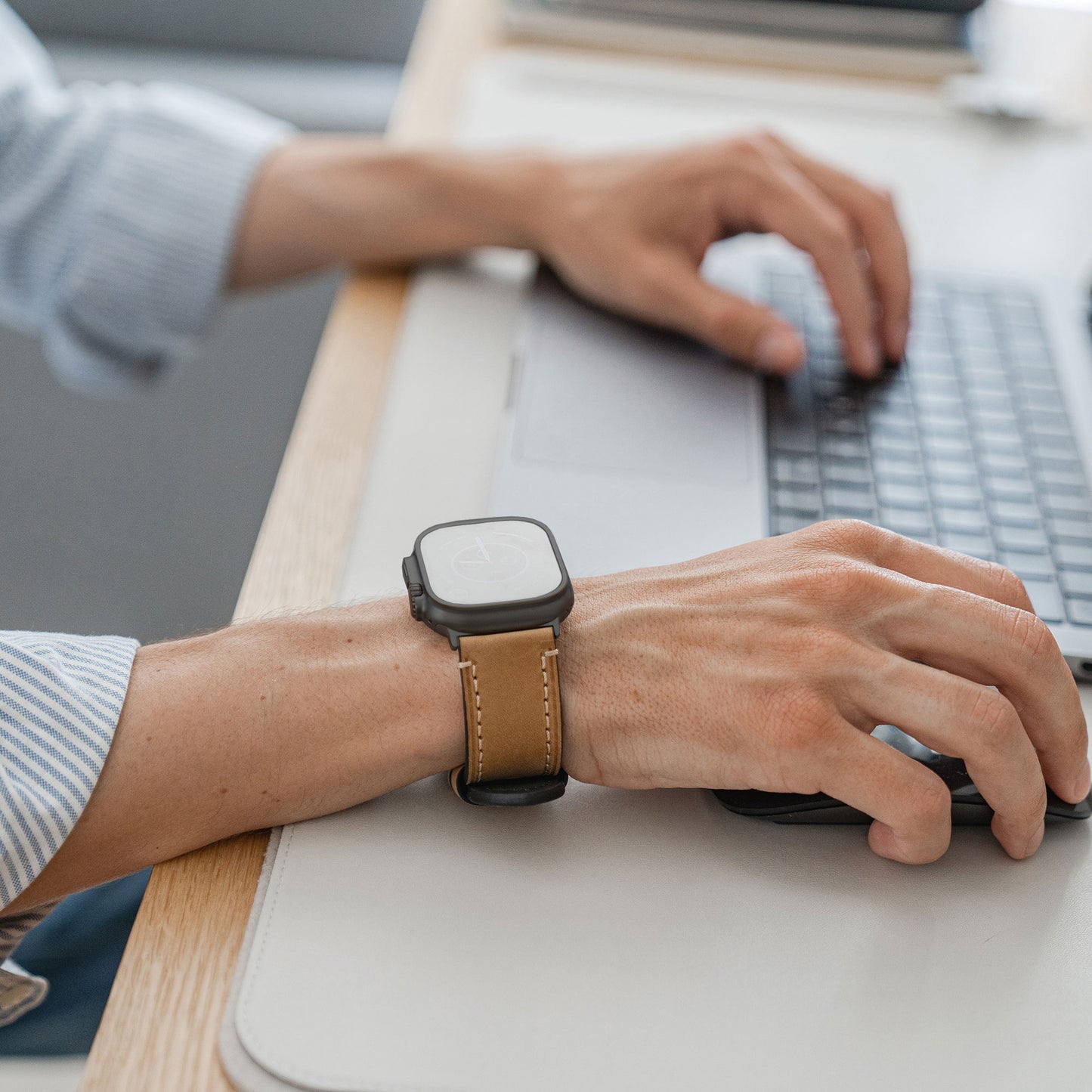 Jemand, der ein Arktisband Apple Watch Ultra PALERMO GRANDE Lederarmband trägt, tippt an einem hellen Schreibtisch auf einem Laptop, wobei nur seine Hände und ein Teil eines gestreiften Hemdärmels sichtbar sind.