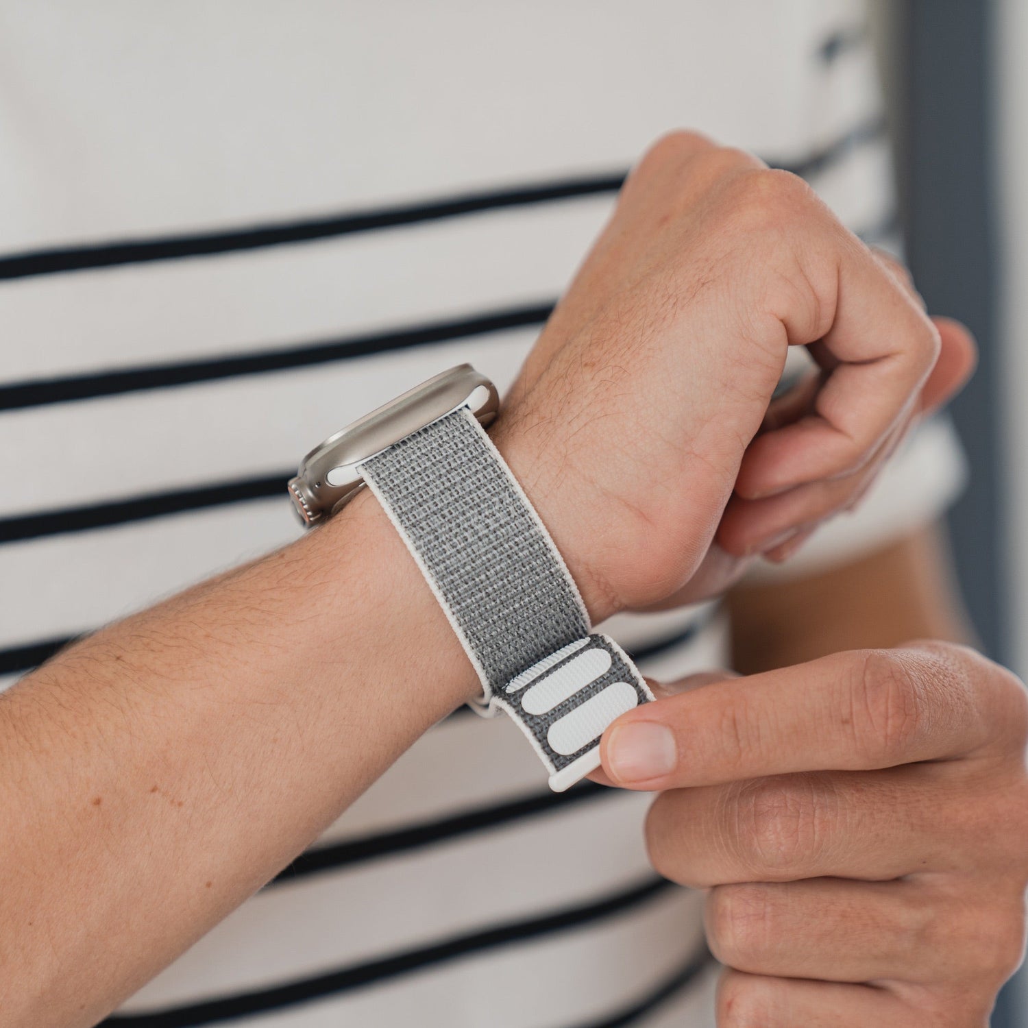 Eine Person befestigt ein graues Smartwatch-Armband an ihrem Handgelenk und trägt ein weiß-schwarz gestreiftes Hemd.
