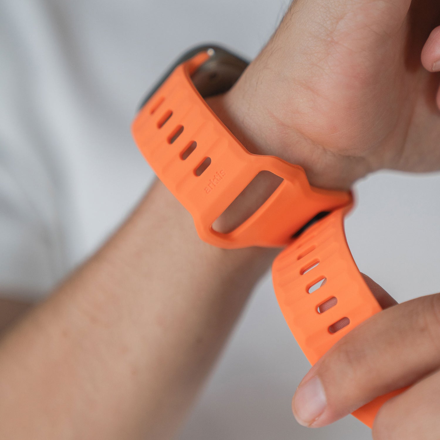 Eine Person befestigt ein orangefarbenes Arktisband Apple Watch Silikonarmband „Active“ um ihr Handgelenk. Ihr Oberkörper im weißen Hemd ist teilweise vor einem unscharfen Hintergrund sichtbar. Das Armband bietet optimale Atmungsaktivität für ganztägigen Tragekomfort.