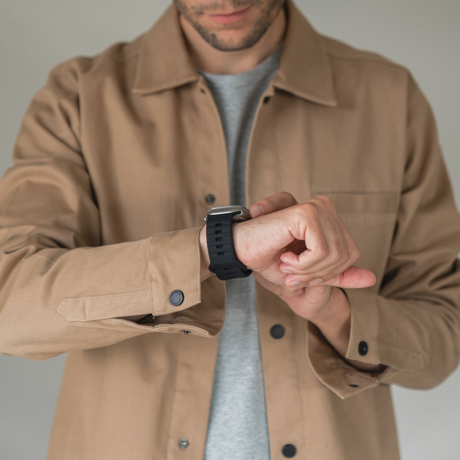 Eine Person in hellbrauner Jacke und grauem Hemd blickt auf ihr linkes Handgelenk und trägt ein arktisband Apple Watch Silikonarmband "Active" von arktisband. Der schlichte Hintergrund ist unscharf.