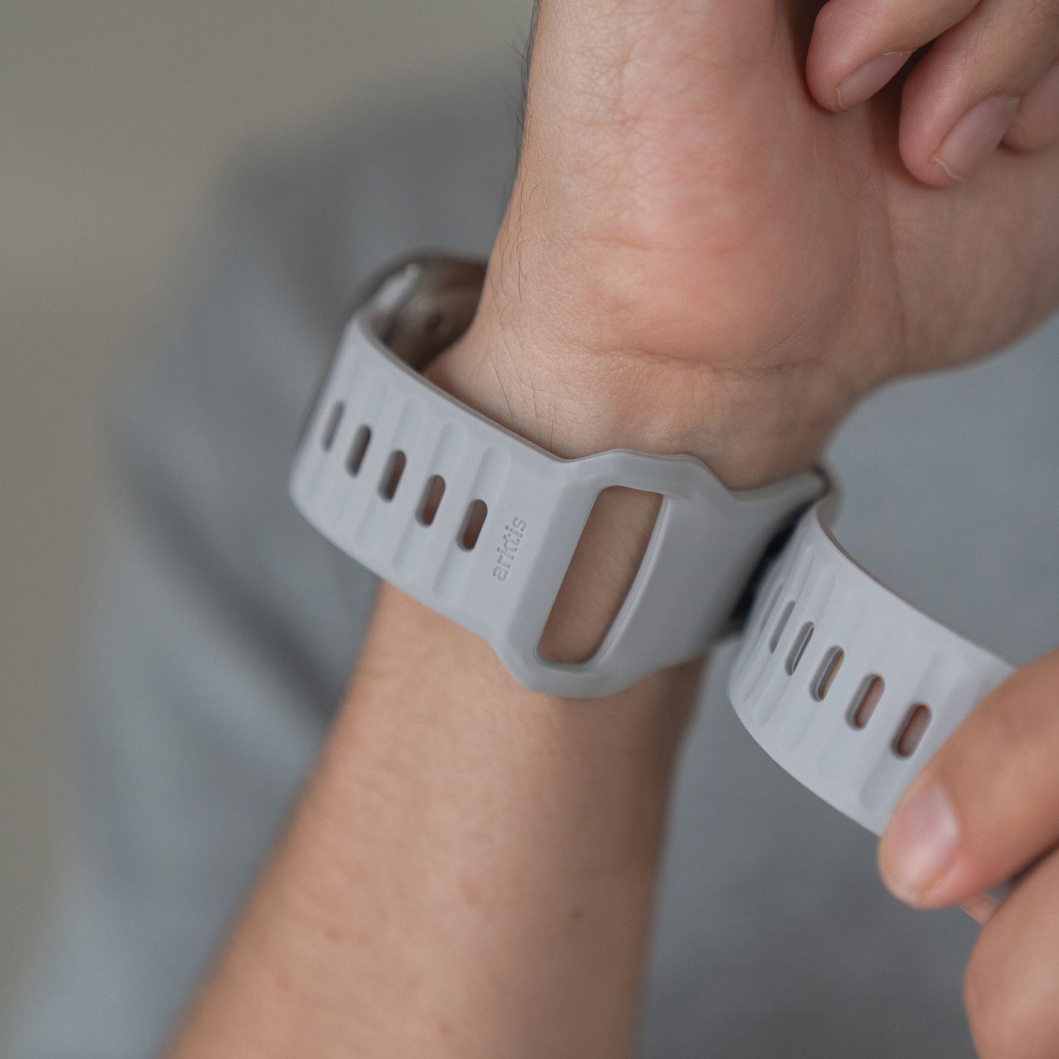 Eine Nahaufnahme einer Person, die ein Arktisband Apple Watch Silikonarmband „Active“ in Grau um ihr Handgelenk legt, wobei das atmungsaktive Design und der Verschluss des Bandes vor einem neutralen, unscharfen Hintergrund hervorgehoben werden.
