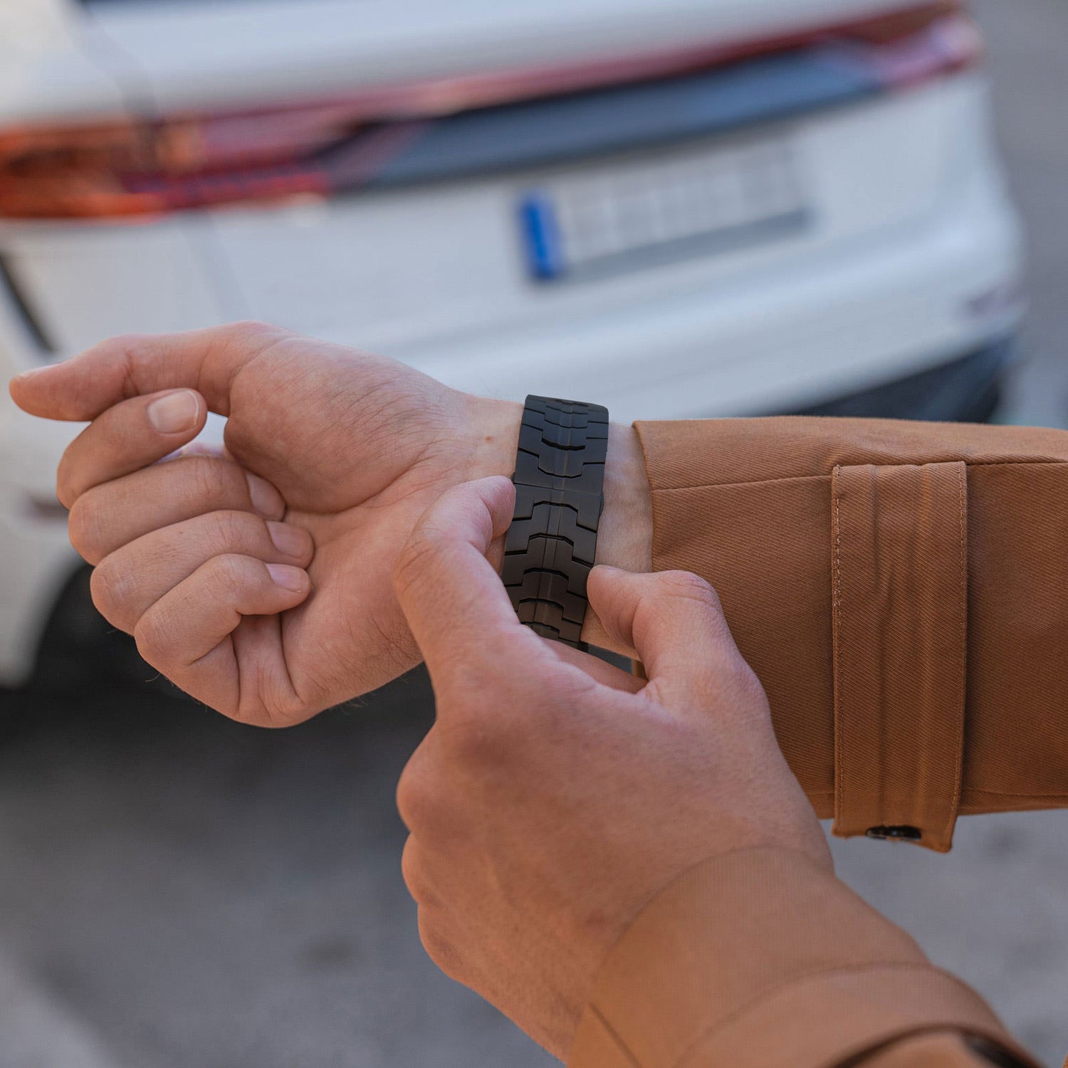 Eine Person in einer braunen Jacke befestigt ein arktisband Apple Watch Ultra Armband TITANIUM F-15 von arktisband an ihrem linken Handgelenk, im Hintergrund ist ein weißes Auto mit einem unscharfen Nummernschild zu sehen.
