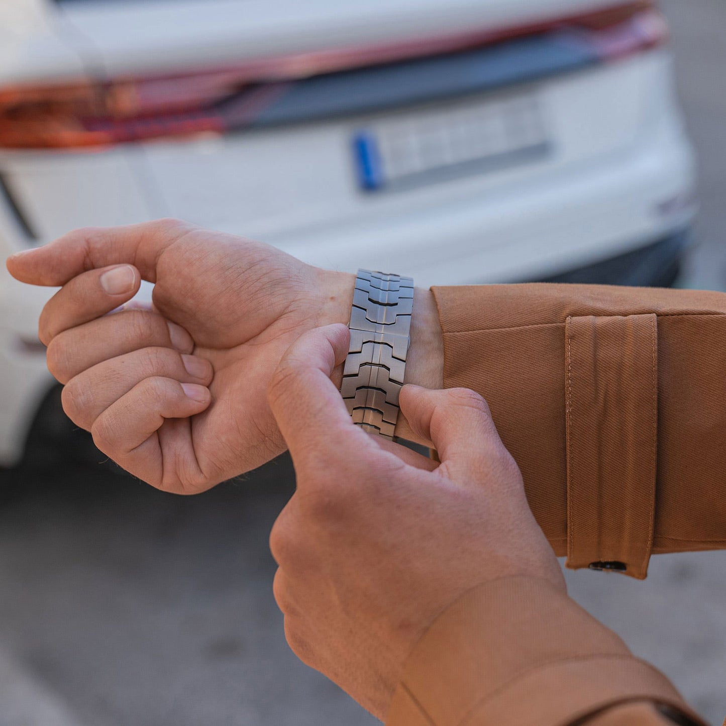 Eine Person in einer braunen Jacke rückt ein Arktisband Apple Watch TITAN Armband „Aviato Edition“ an ihrem Handgelenk zurecht und steht vor einem weißen Auto mit verschwommenem Nummernschild.