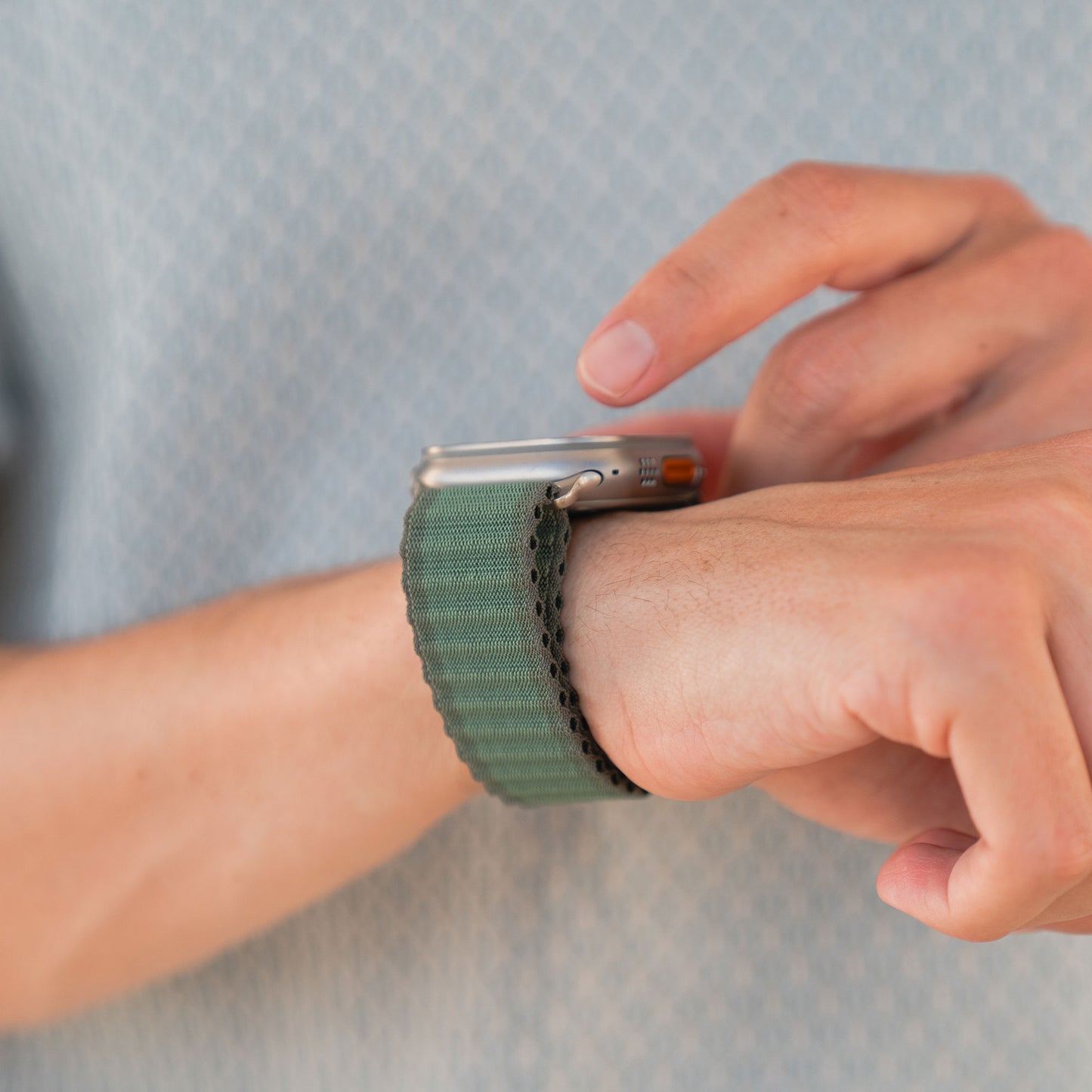 Eine Person, die ein Arktisband Apple Watch Nylonarmband „Venture“ mit grünem Stoffband trägt, berührt den Bildschirm vor einem verschwommenen hellblauen Hemd – ideal für Outdoor-Enthusiasten, die sowohl Wert auf Stil als auch auf Funktionalität legen.