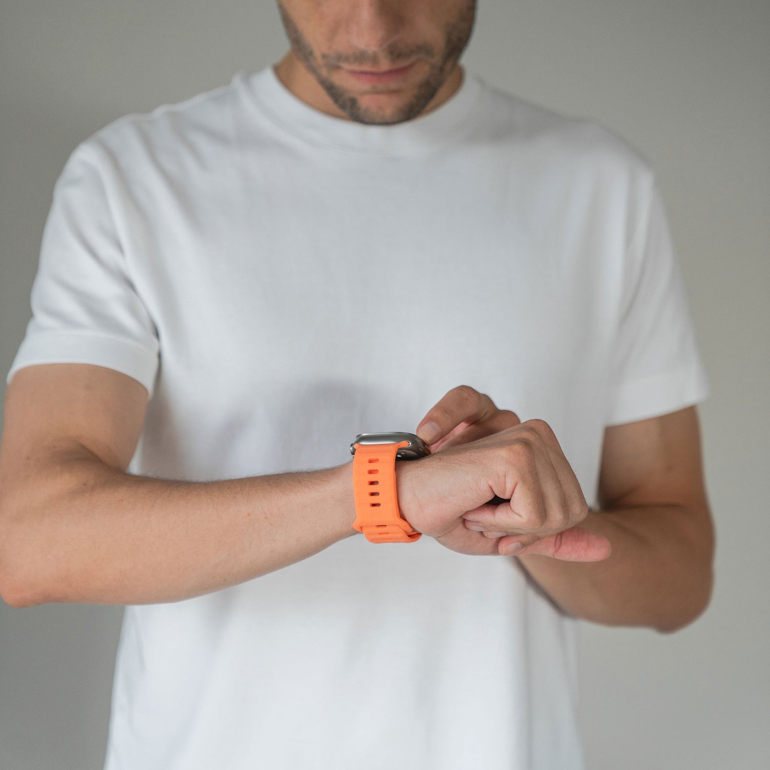 Jemand trägt ein schlichtes weißes T-Shirt und prüft sein arktisband Apple Watch Silikonarmband „Active“ in Orange von arktisband an seinem linken Handgelenk vor einem neutralen grauen Hintergrund.