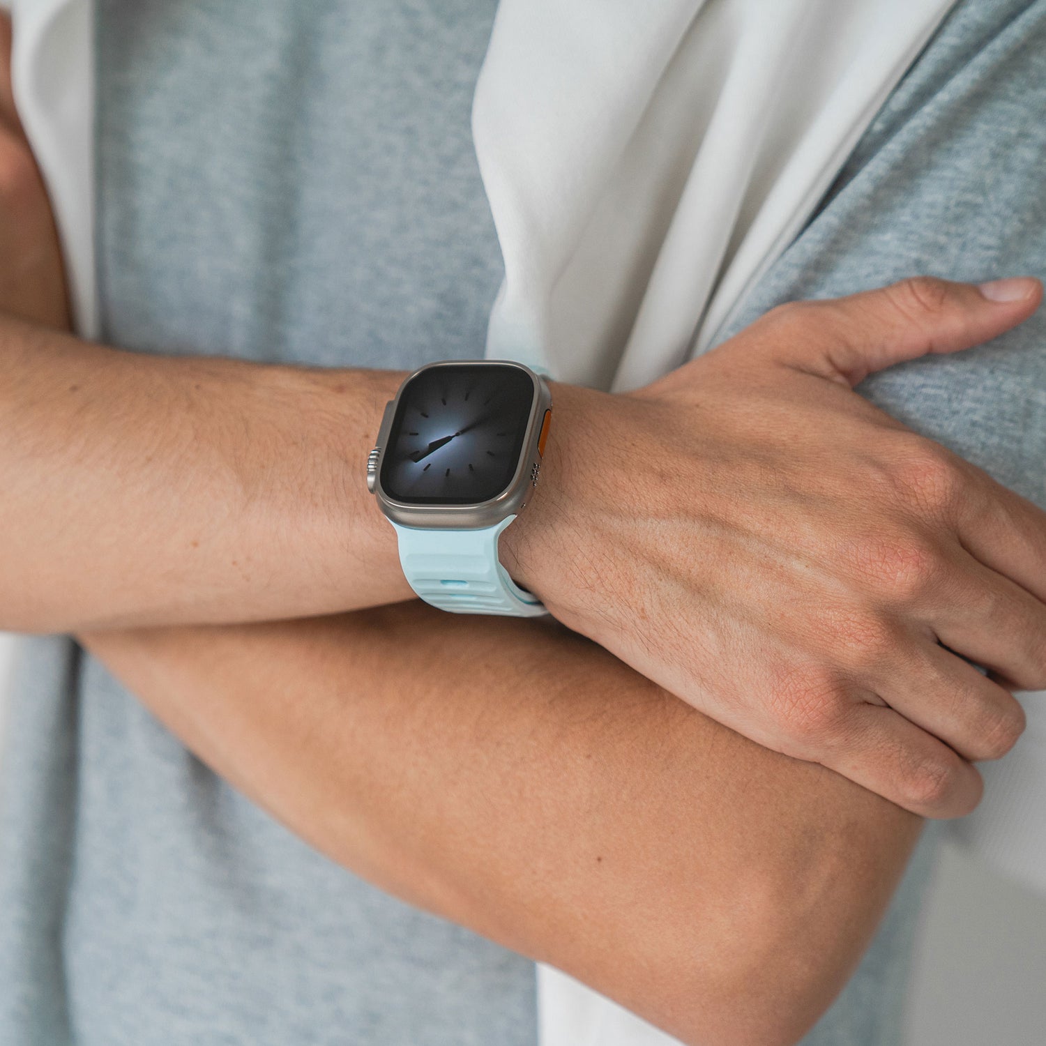 Eine Person mit verschränkten Armen trägt ein hellgraues Hemd und ein Arktisband Apple Watch Silikonarmband „Active“ in Hellblau am linken Handgelenk. Das atmungsaktive Design sorgt für ganztägigen Tragekomfort. Der Bildschirm der Smartwatch ist schwarz und leer.