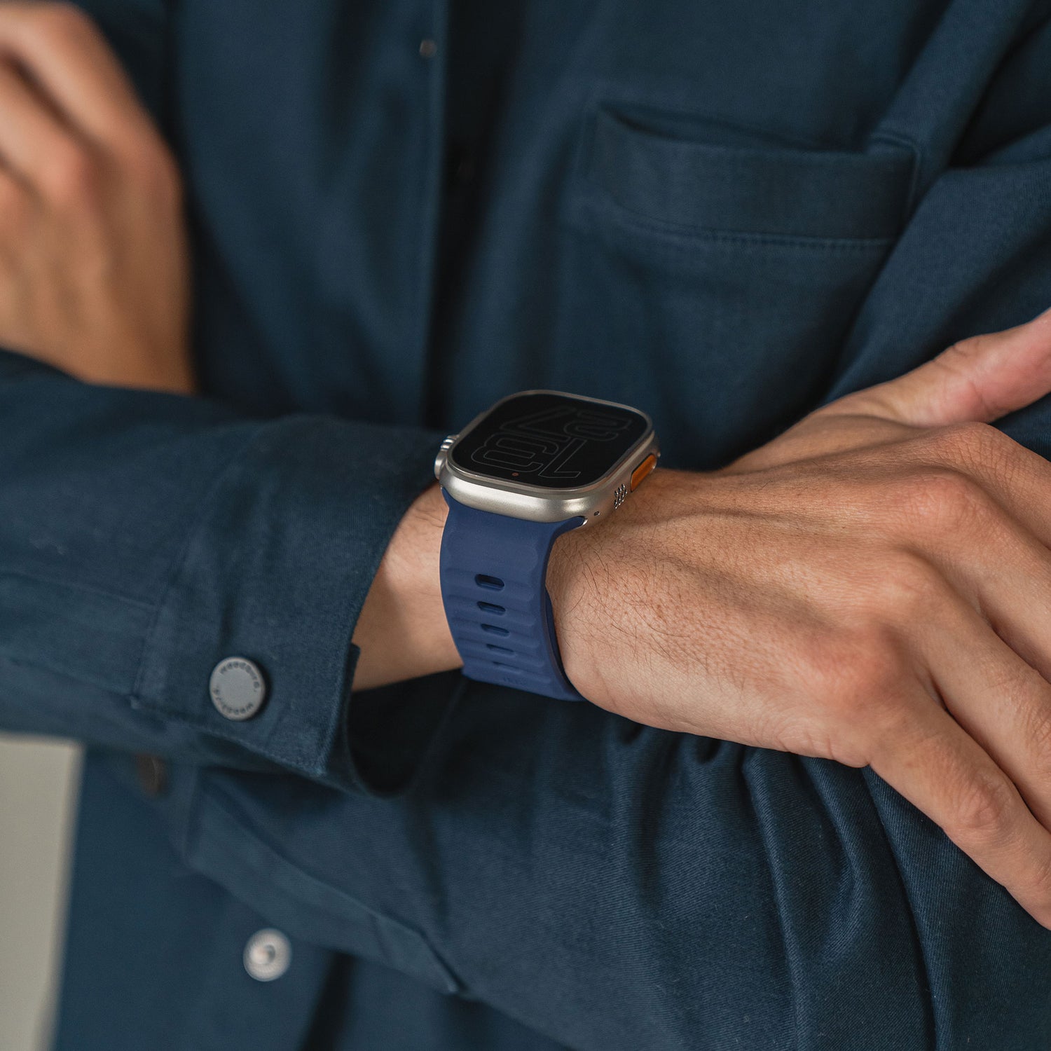 Eine Person in einem dunkelblauen Hemd verschränkt die Arme und zeigt an ihrem linken Handgelenk ein Arktisband Apple Watch Silikonarmband „Active“ mit blauem Band und großem, quadratischem Zifferblatt.