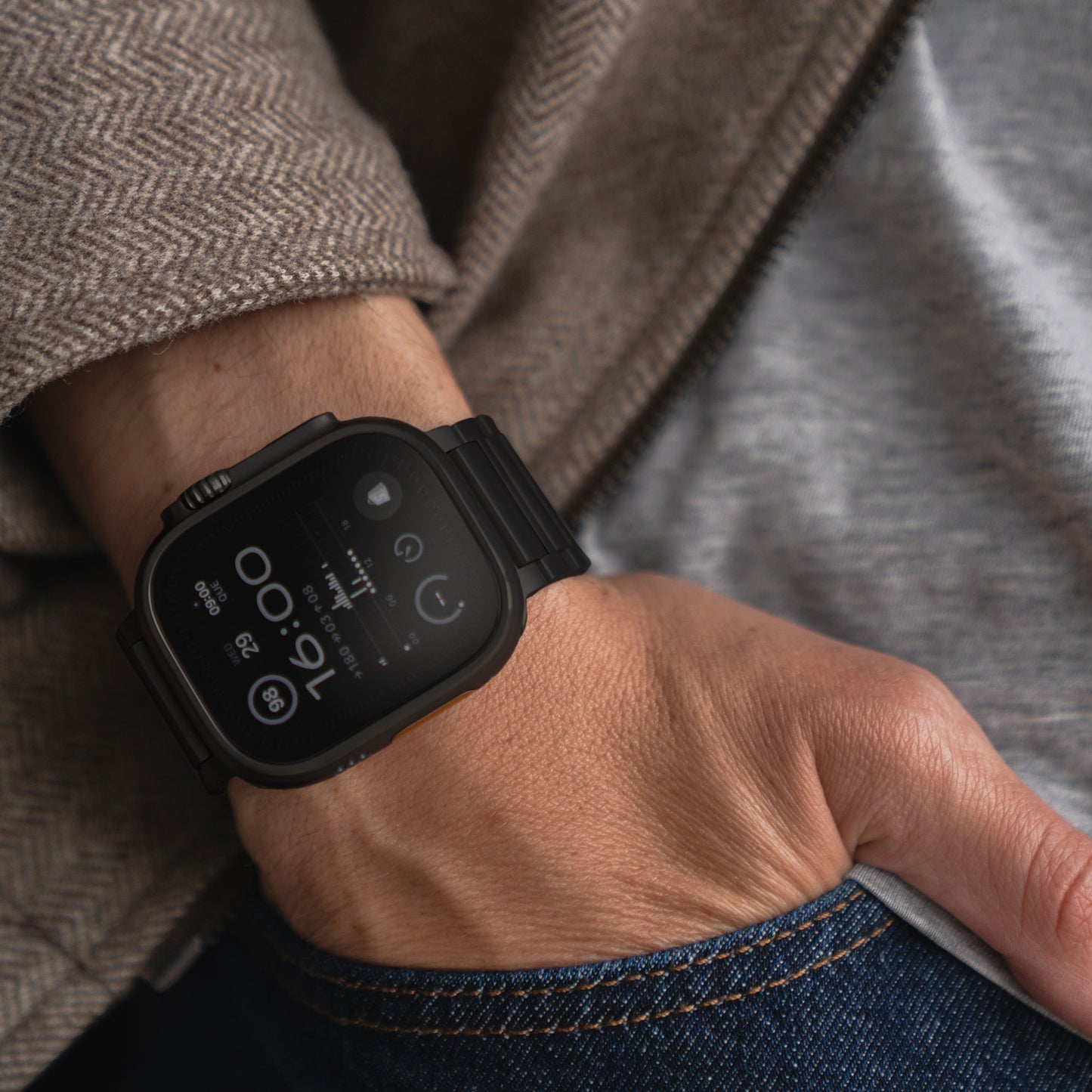 Eine Person trägt ein arktisband Apple Watch Ultra Titanarmband „Maverick“ von arktisband am linken Handgelenk, kombiniert mit einer grau-braunen Fischgrätjacke und Bluejeans. Die Uhr zeigt Uhrzeit, Datum, Wetter und Batteriestatus an.