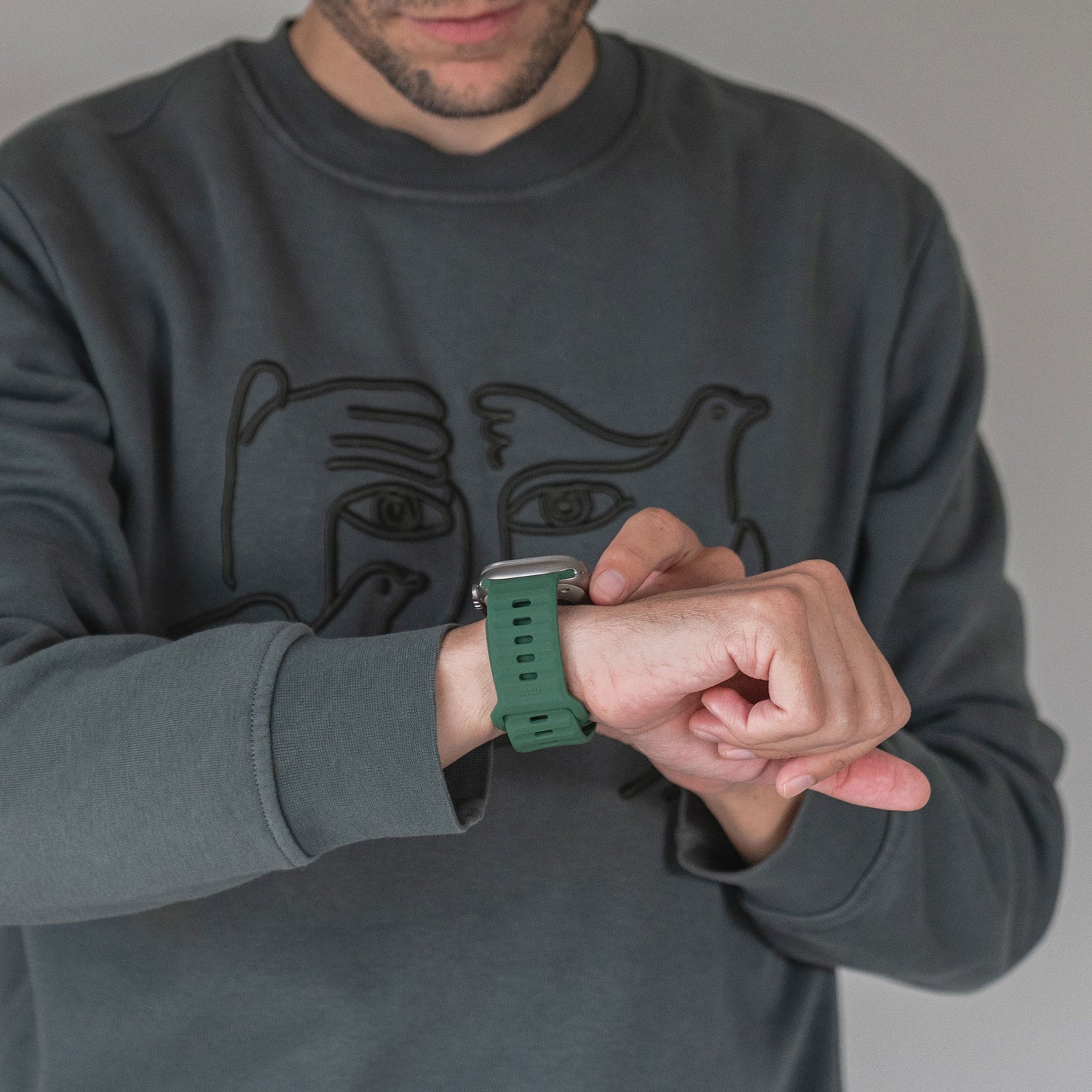 Eine Person in einem grauen Sweatshirt mit Gesichts- und Vogelmotiv rückt ein grünes Arktisband Apple Watch Silikonarmband „Active“ an ihrem linken Handgelenk zurecht, wobei ihr Gesicht teilweise außerhalb des Bildes liegt.
