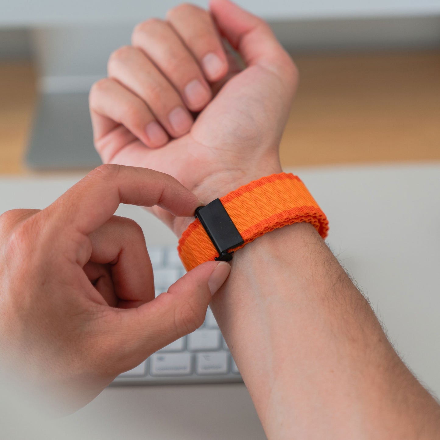 Jemand befestigt das orangefarbene Arktisband Apple Watch Nylonarmband „Venture“ mit schwarzem Verschluss um sein Handgelenk. Eine Computertastatur im Hintergrund lässt vermuten, dass sich dieses Arktisband sowohl für die Arbeit als auch für Outdoor-Aktivitäten eignet.