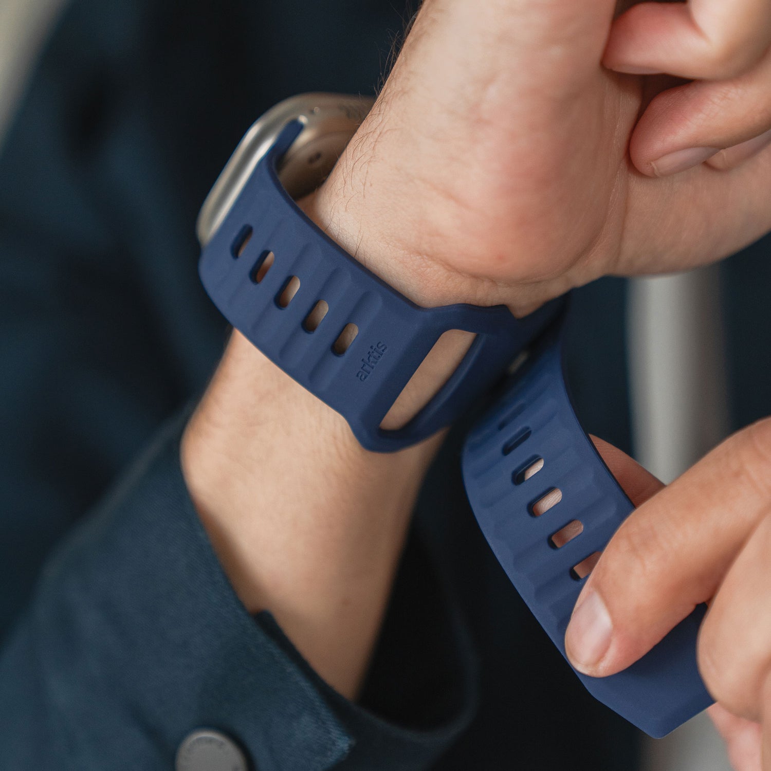 Eine Person in einem blauen Ärmel mit Knöpfen befestigt das arktisband Apple Watch Silikonarmband „Active“ mit atmungsaktivem Design von arktisband an ihrem Handgelenk, wobei das Zifferblatt der Apple Watch teilweise sichtbar ist.