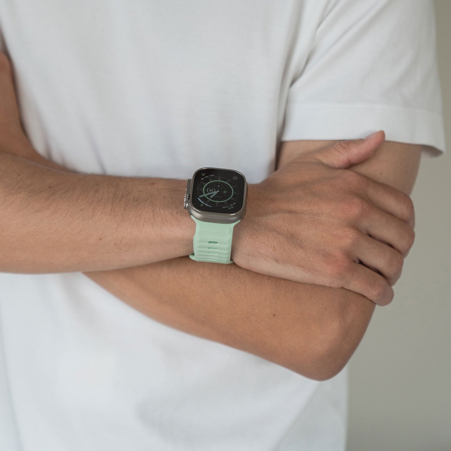 Eine Person in einem weißen Kurzarm-T-Shirt verschränkt die Arme und präsentiert ein arktisband Apple Watch Silikonarmband „Active“ in Hellgrün an ihrem linken Handgelenk vor einem schlichten, neutralen Hintergrund für einen sportlichen Look.