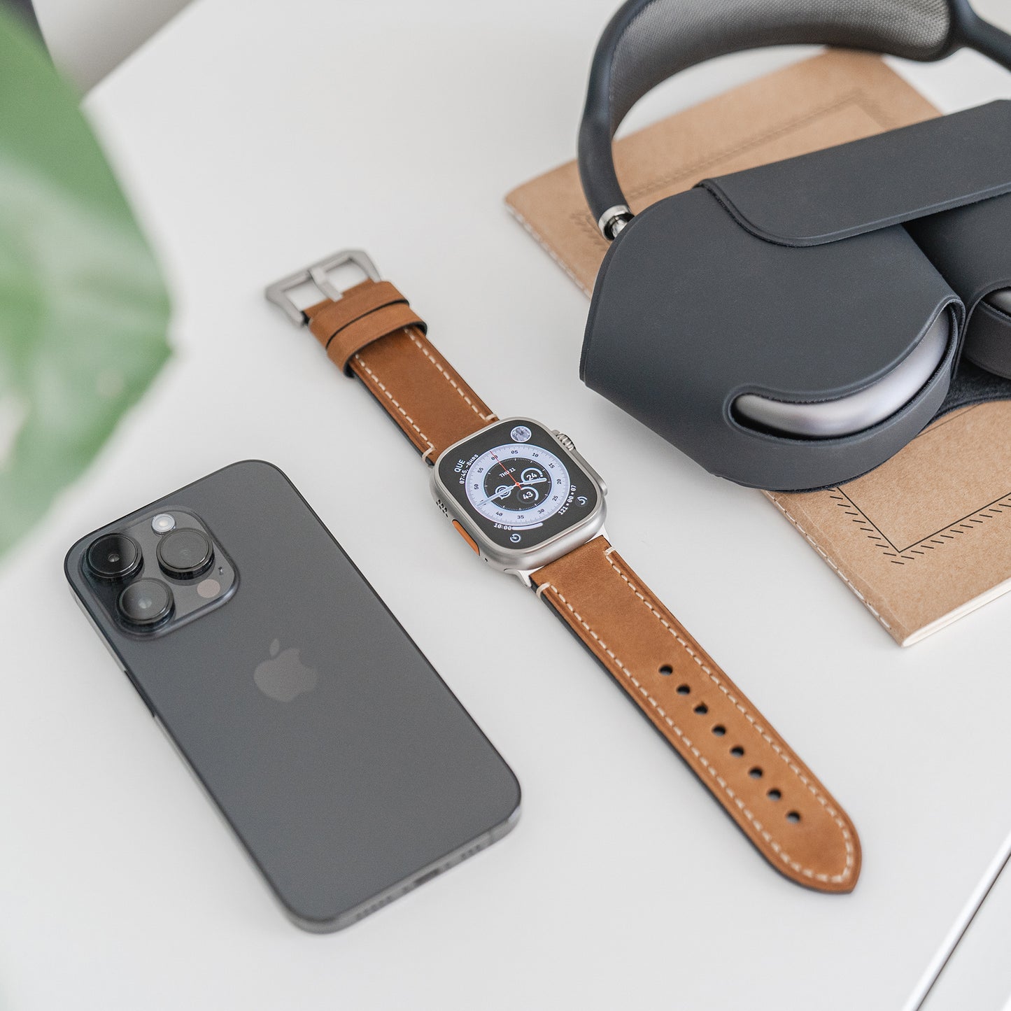 Eine Arktisband Apple Watch Ultra mit einem PALERMO GRANDE Lederarmband, ein schwarzes Smartphone und Over-Ear-Kopfhörer liegen auf einem geschlossenen Notebook auf einer weißen Fläche. Im Vordergrund ist ein unscharfes grünes Objekt zu sehen.