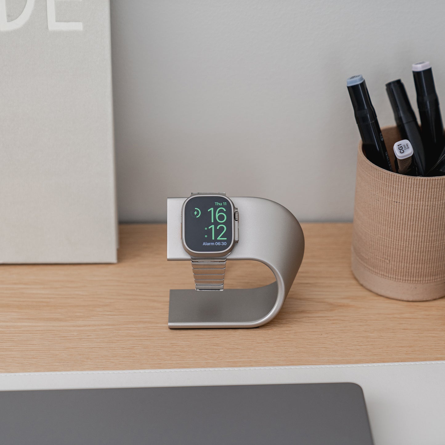 Ein Arktisband Aluminium Apple Watch Ultra Ständer in Titanfarben hält eine Apple Watch Ultra elegant neben Stiften auf einem aufgeräumten Holzschreibtisch.