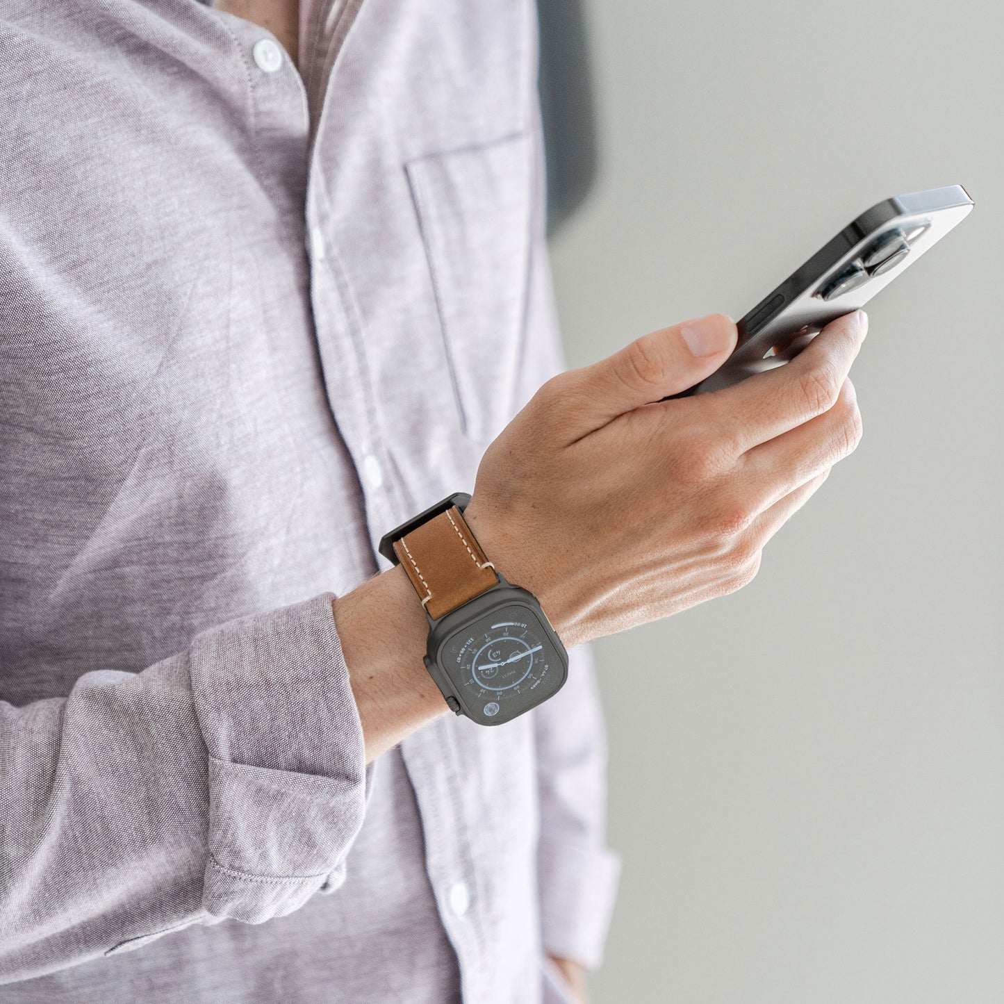 Eine Person in einem lavendelfarbenen Hemd hält ein Smartphone und trägt eine Arktisband Apple Watch Ultra mit dem PALERMO GRANDE Lederarmband, akzentuiert mit 316L-Edelstahl, auf dessen Handgelenk ein digitales Zifferblatt zu sehen ist.