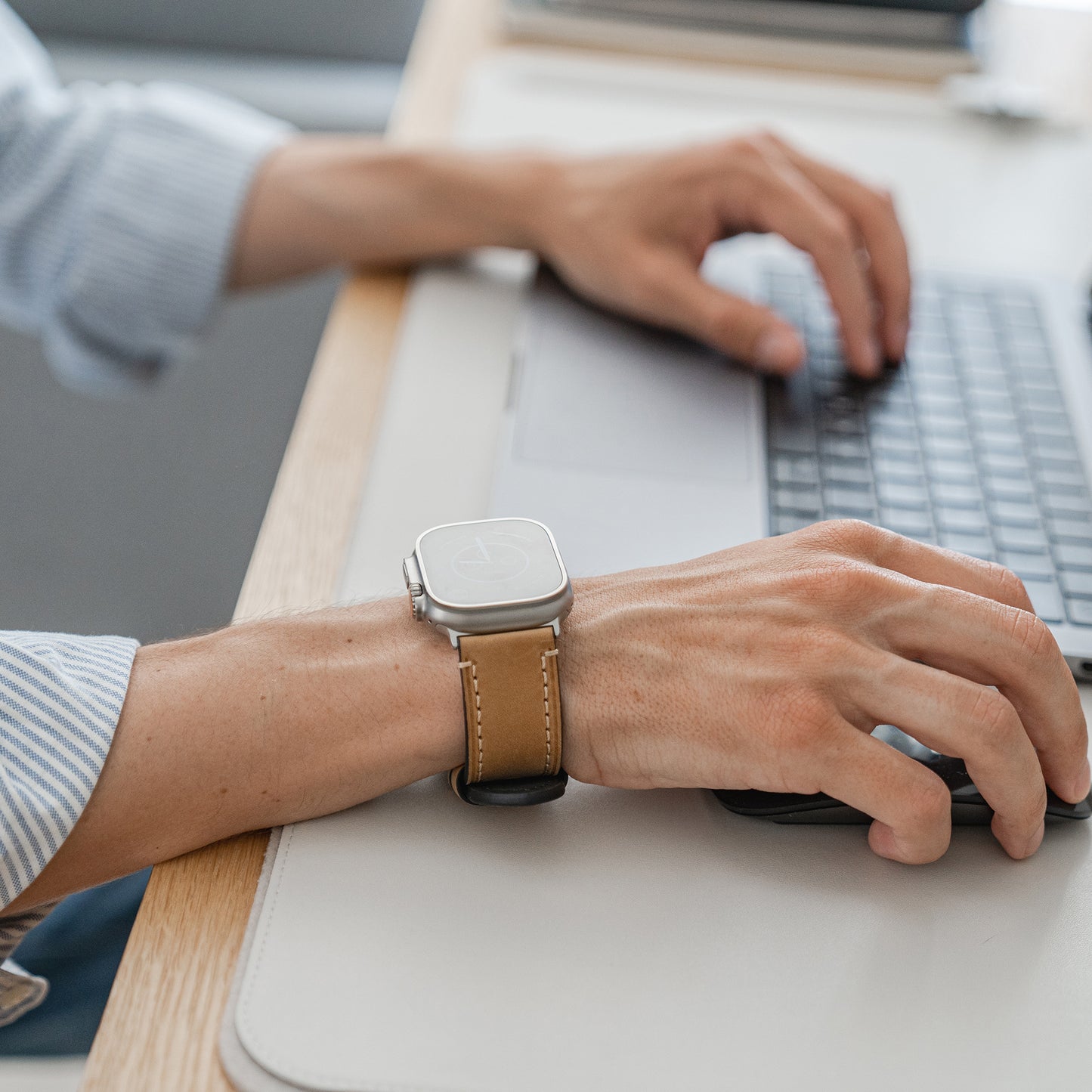 Jemand trägt ein Arktisband Apple Watch Ultra PALERMO GRANDE Lederarmband und tippt auf einem Laptop, sein linker Arm ruht auf einer hellen Schreibtischunterlage.