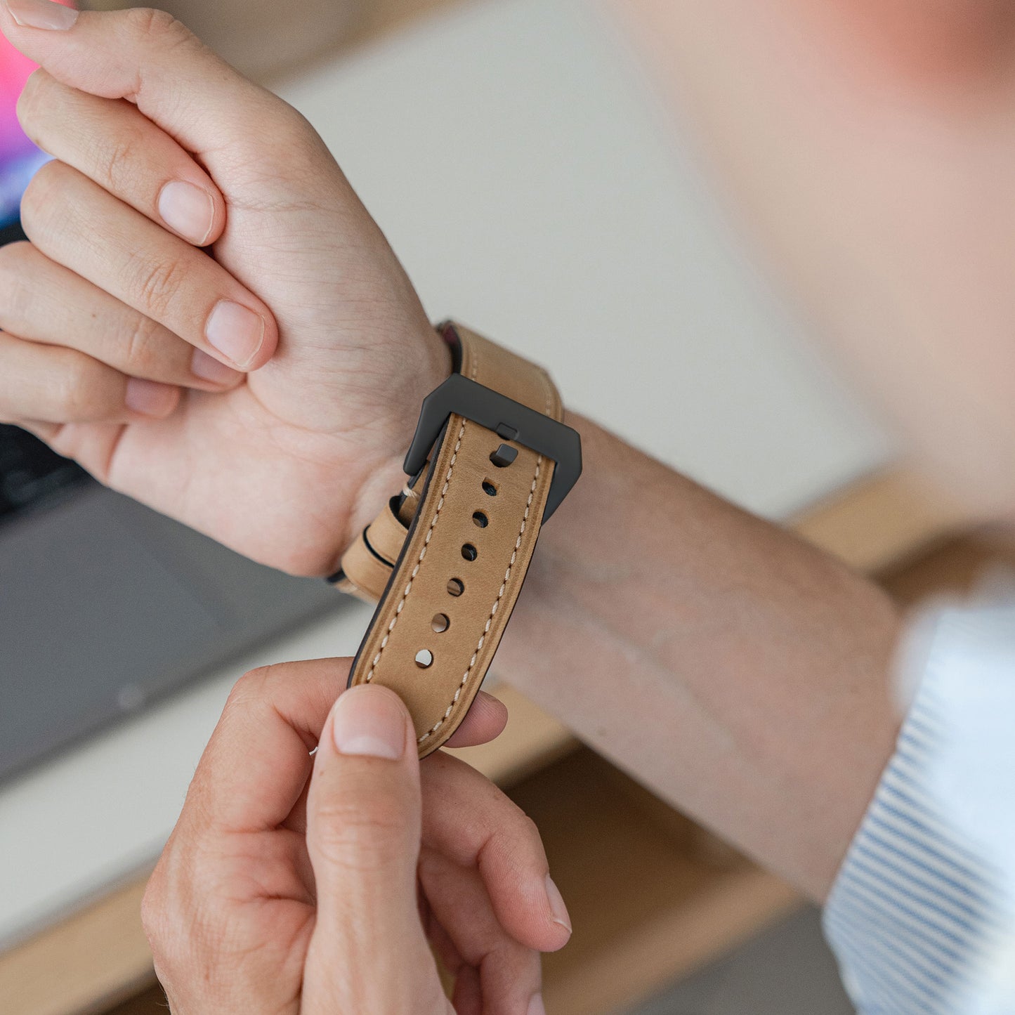 Jemand befestigt ein arktisband Apple Watch Ultra PALERMO GRANDE Lederarmband von arktisband mit einer schwarzen Schnalle aus 316L-Edelstahl um sein Handgelenk, während im Hintergrund ein Teil eines Laptops sichtbar ist.