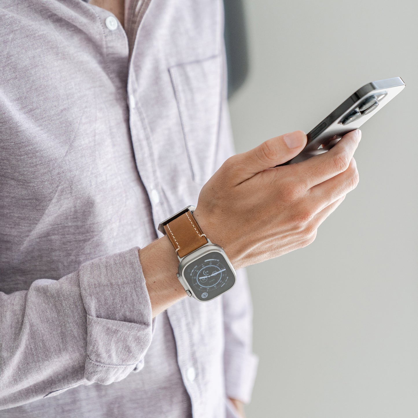 Eine Person in einem hellvioletten Hemd hält ein Smartphone und trägt ein arktisband Apple Watch Ultra PALERMO GRANDE Lederarmband am Handgelenk. Der Hintergrund ist schlicht und neutral.