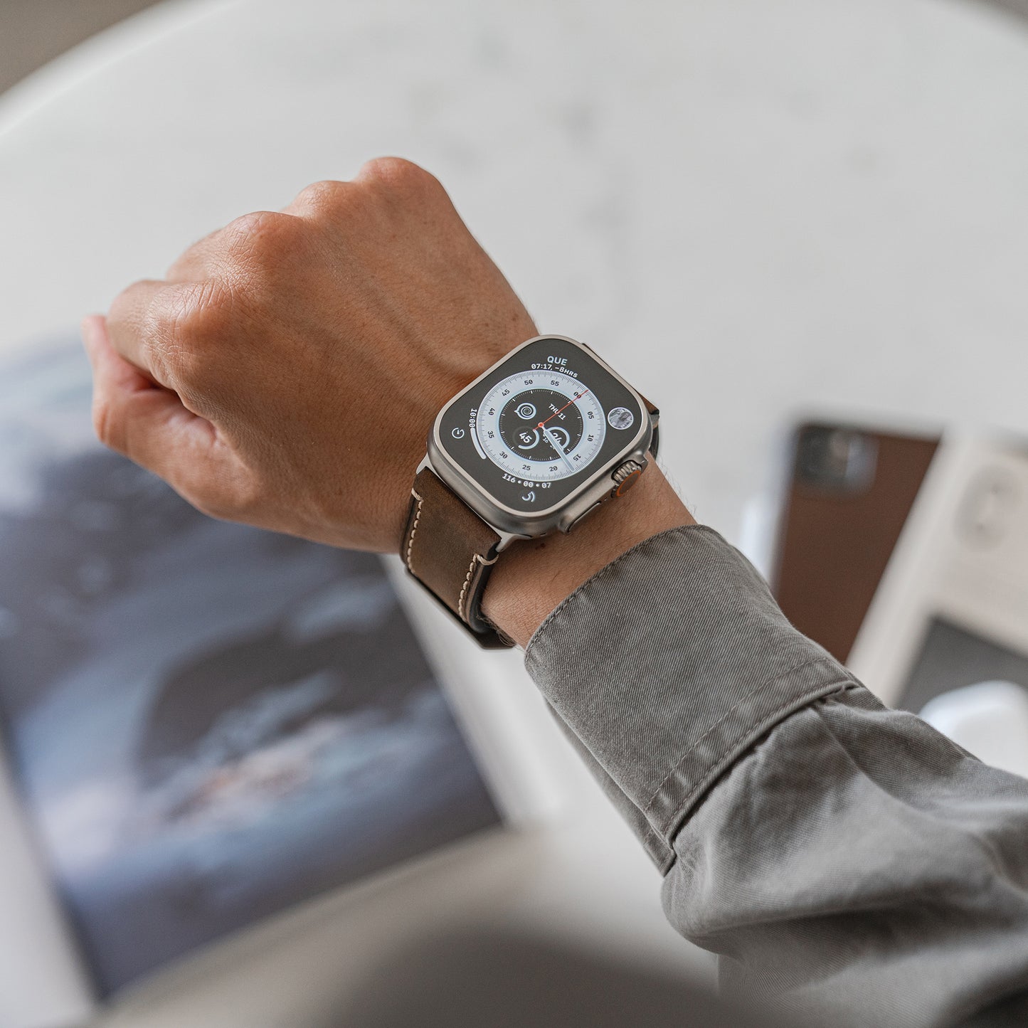 Eine Person, die ein Arktisband Apple Watch Ultra PALERMO GRANDE Lederarmband trägt, überprüft das Zifferblatt. Im Hintergrund liegen eine offene Zeitschrift, ein Smartphone und ein weiteres elektronisches Gerät auf einem weißen Tisch.