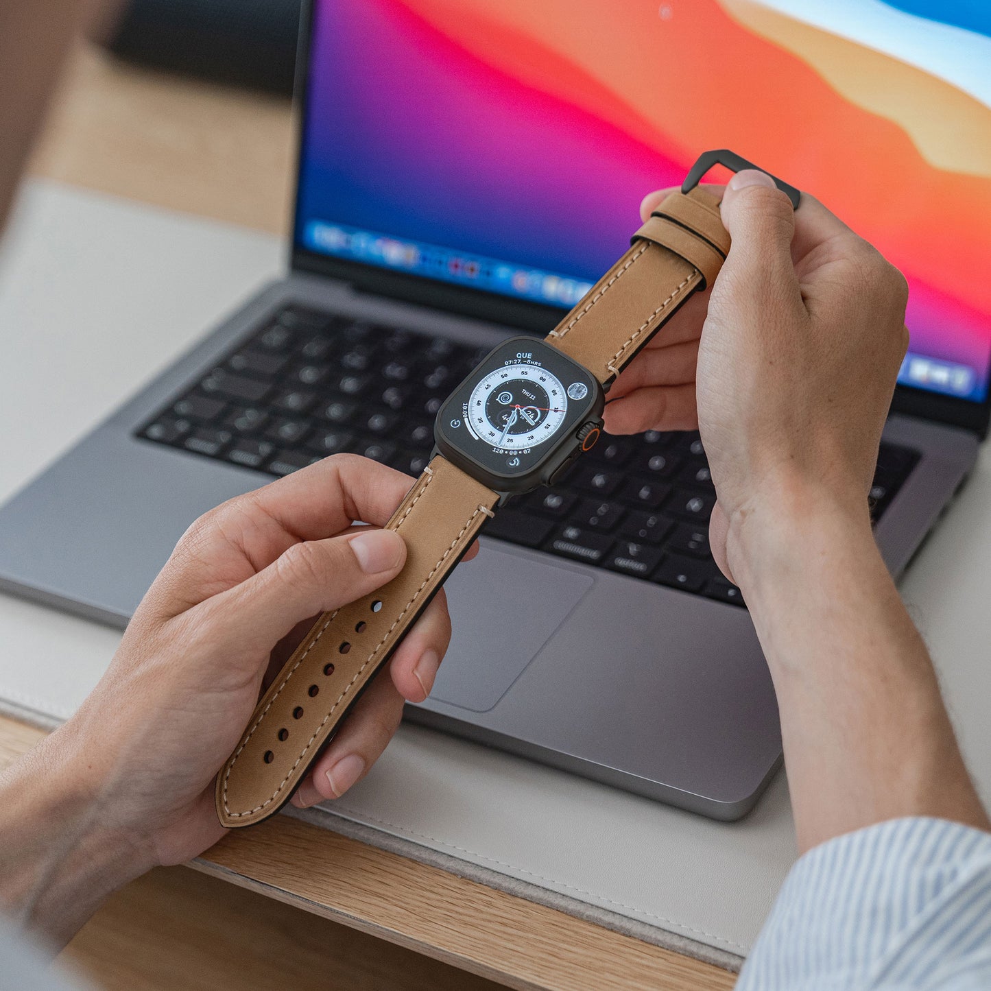 Eine Person hält eine Apple Watch Ultra mit Arktisband, PALERMO GRANDE Lederarmband und 316L Edelstahl-Akzenten vor einem Laptop mit buntem Bildschirm. Die Uhr hat ein analoges Zifferblatt mit mehreren Zifferblättern.