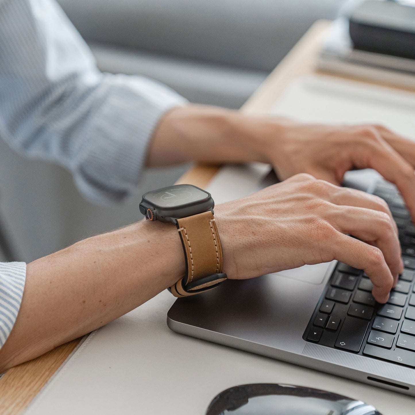 Eine Person trägt ein Arktisband Apple Watch Ultra PALERMO GRANDE Lederarmband mit 316L-Edelstahlverschluss und tippt an einem Schreibtisch auf einem Laptop, bekleidet mit einem hellblauen, gestreiften Hemd.