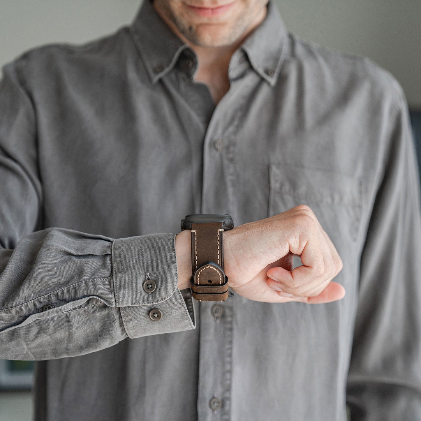 Eine Person in einem grauen Hemd mit Knöpfen blickt auf ihr Handgelenk, an dem ein Arktisband Apple Watch Ultra PALERMO GRANDE Lederarmband angebracht ist – ohne Zifferblatt.