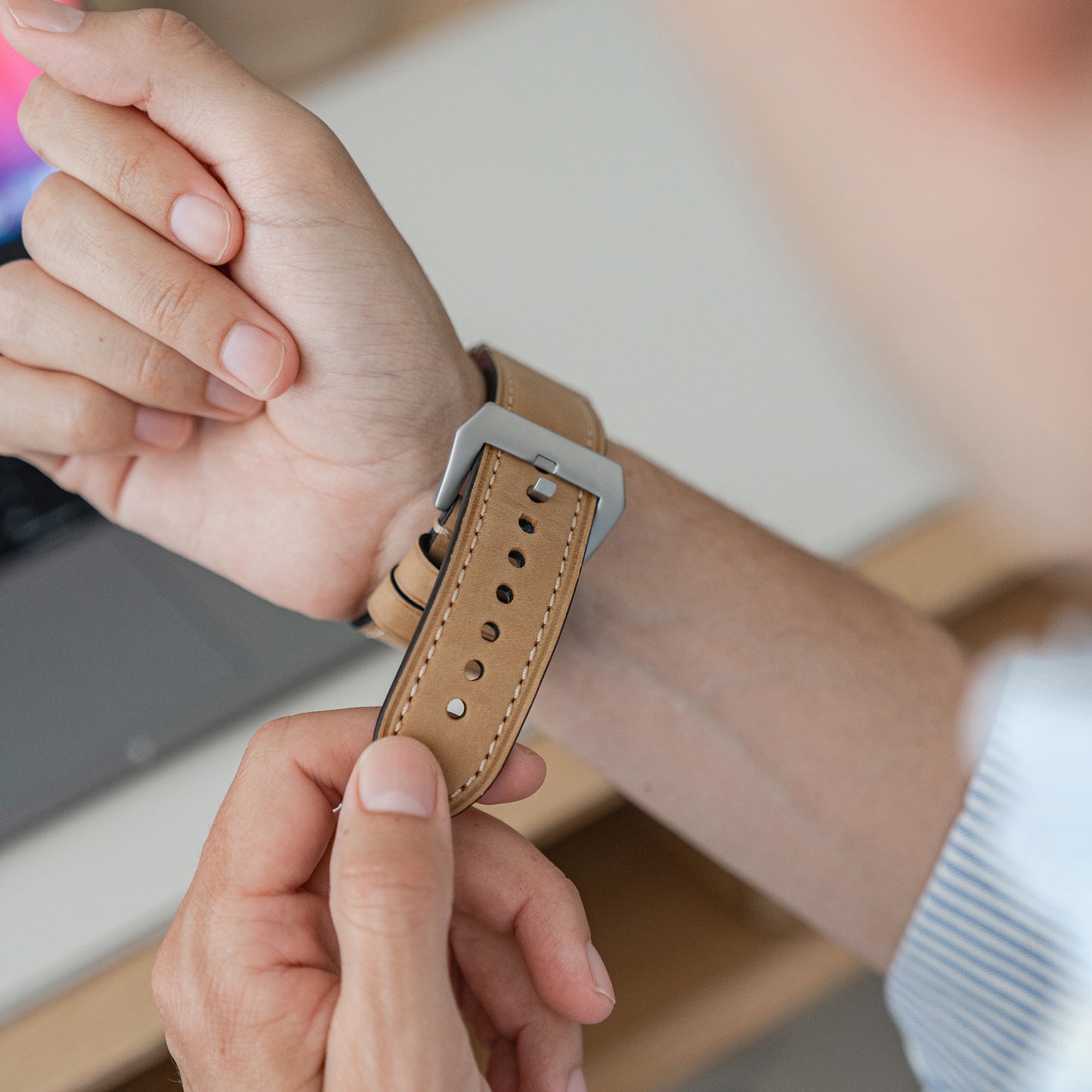 Eine Person trägt ein Arktisband Apple Watch Ultra PALERMO GRANDE Lederarmband, gepaart mit ihrer Apple Watch Ultra. Im Hintergrund sind ein Laptop und ein Holzschreibtisch zu sehen. Die Person trägt ein hell gestreiftes Hemd.