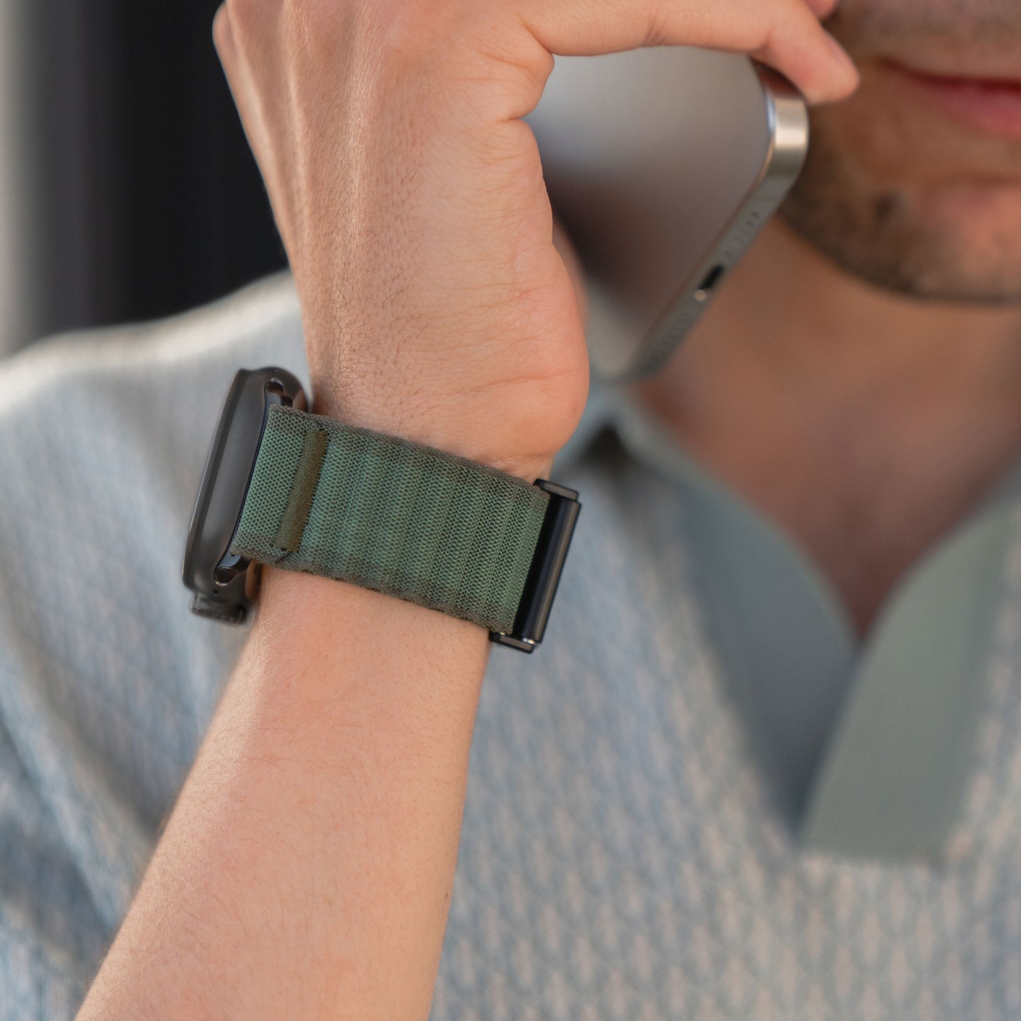 Eine Person mit einem grünen Arktisband Apple Watch Nylonarmband „Venture“ hält ein Smartphone vor ihr Gesicht, wobei nur die untere Hälfte sichtbar ist, dazu ein hellblaues gemustertes Hemd – die perfekte Style-Inspiration für Outdoor-Fans.