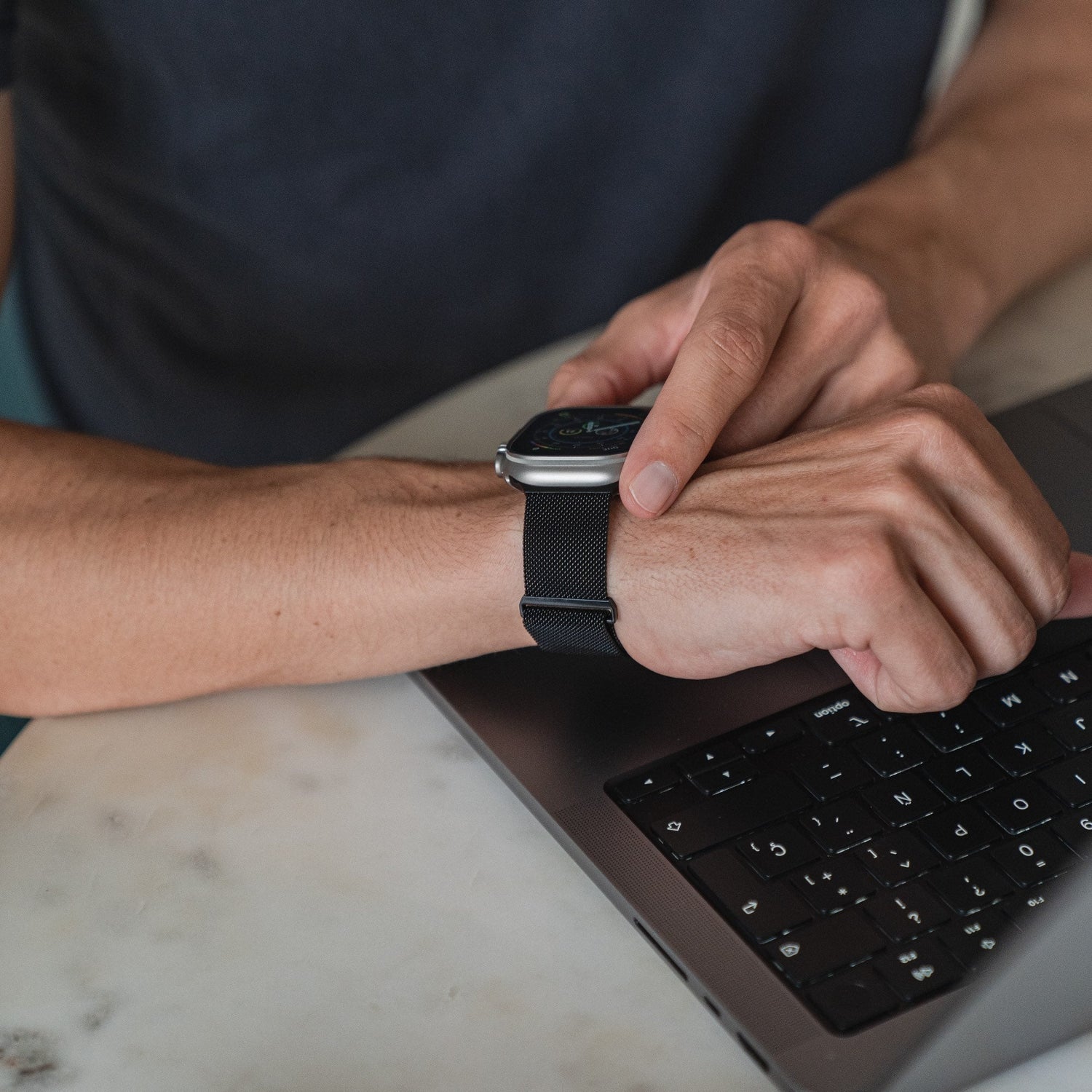 Eine Person trägt ein Arktisband Apple Watch Milanaise Loop Armband und passt es mit der rechten Hand an, während sie einen Laptop auf einem hellen Tisch benutzt. Nur ihre Arme und ein Teil ihres Oberkörpers sind sichtbar.