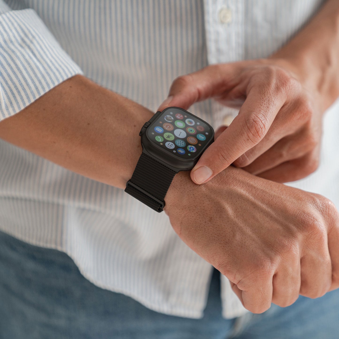 Eine Person in gestreiftem Hemd und Jeans trägt das arktisband Apple Watch Ultra Milanaise Armband. Die Smartwatch von arktisband zeigt mehrere App-Symbole in einem Raster auf ihrem Display an.